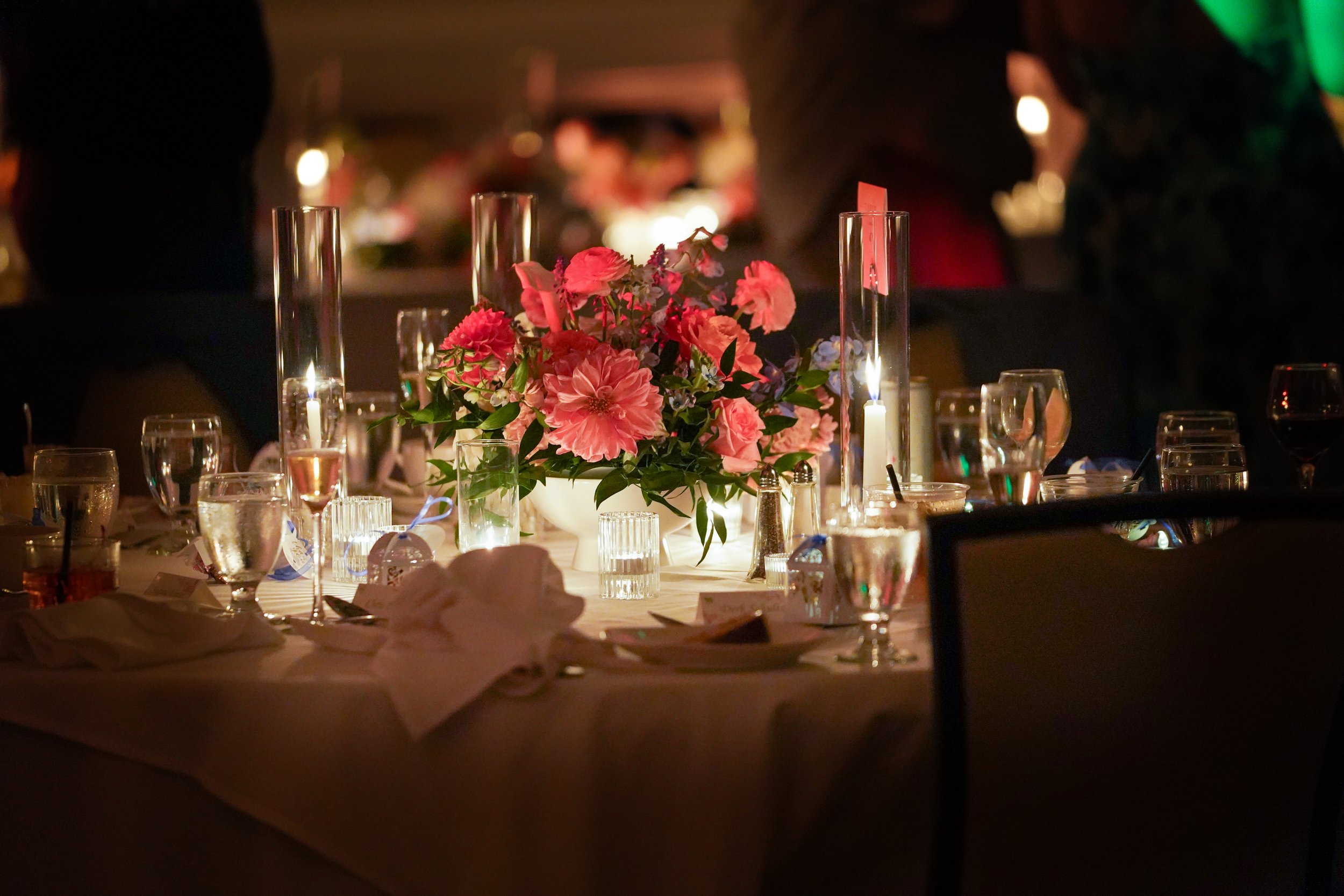 An elegant indoor dining table setting with a pink and blue floral centerpiece, tall glass candle holders, multiple glasses, plates, and white napkins under warm event lighting.