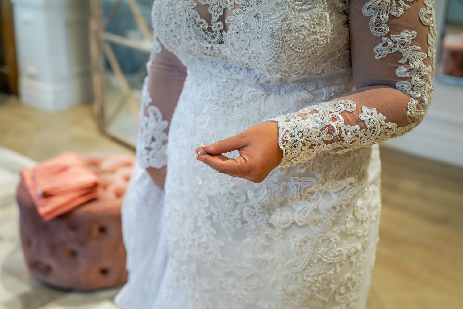 A close-up photograph of a person wearing an ornate white lace wedding dress with long, sheer lace sleeves featuring intricate embroidery and beadwork. The person's right hand is held slightly open near the waist of the dress.