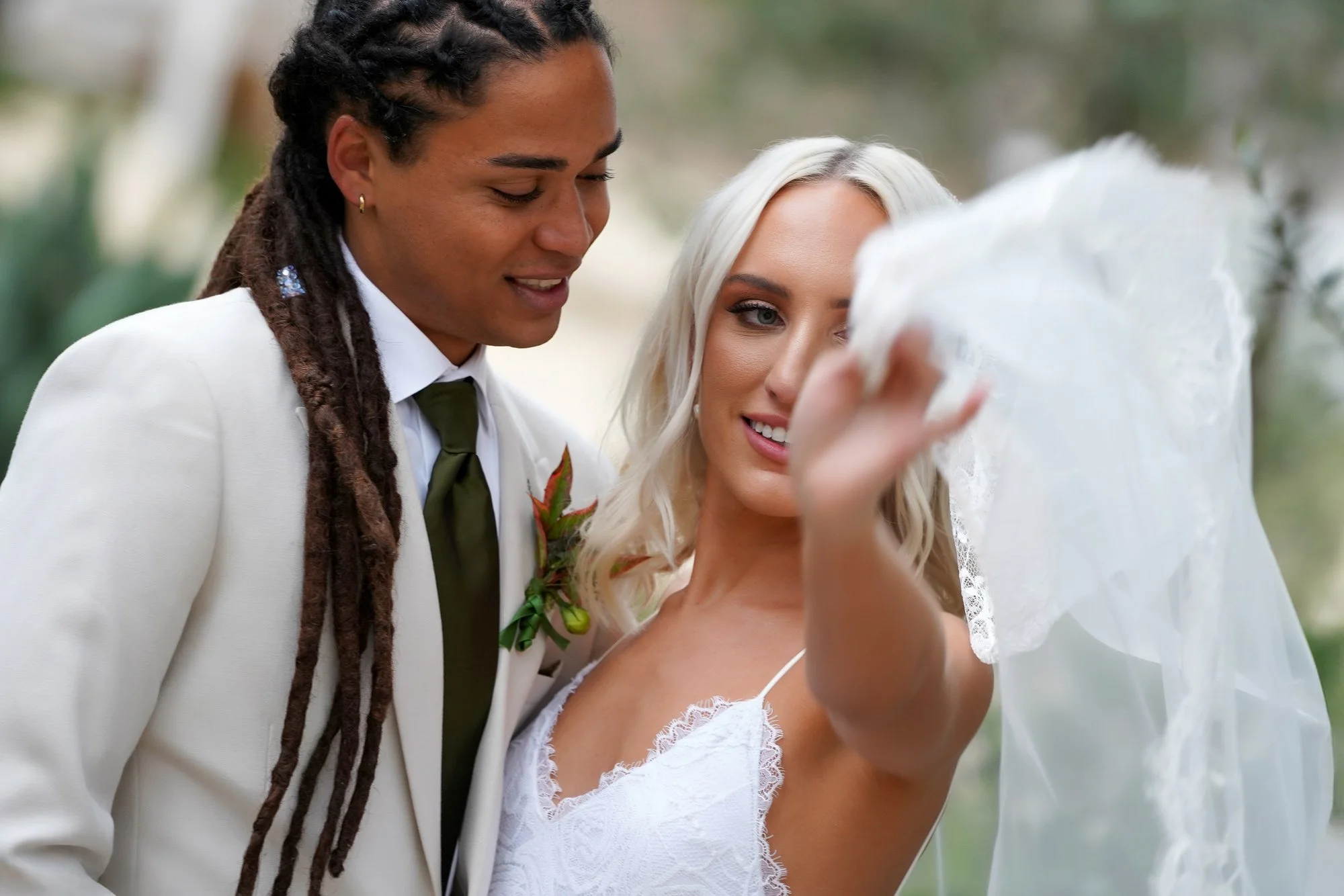 A close-up photo of a newly married couple posing for a portrait, with one wearing a cream-colored suit and the other in a white lace slip dress and veil, both smiling happily.