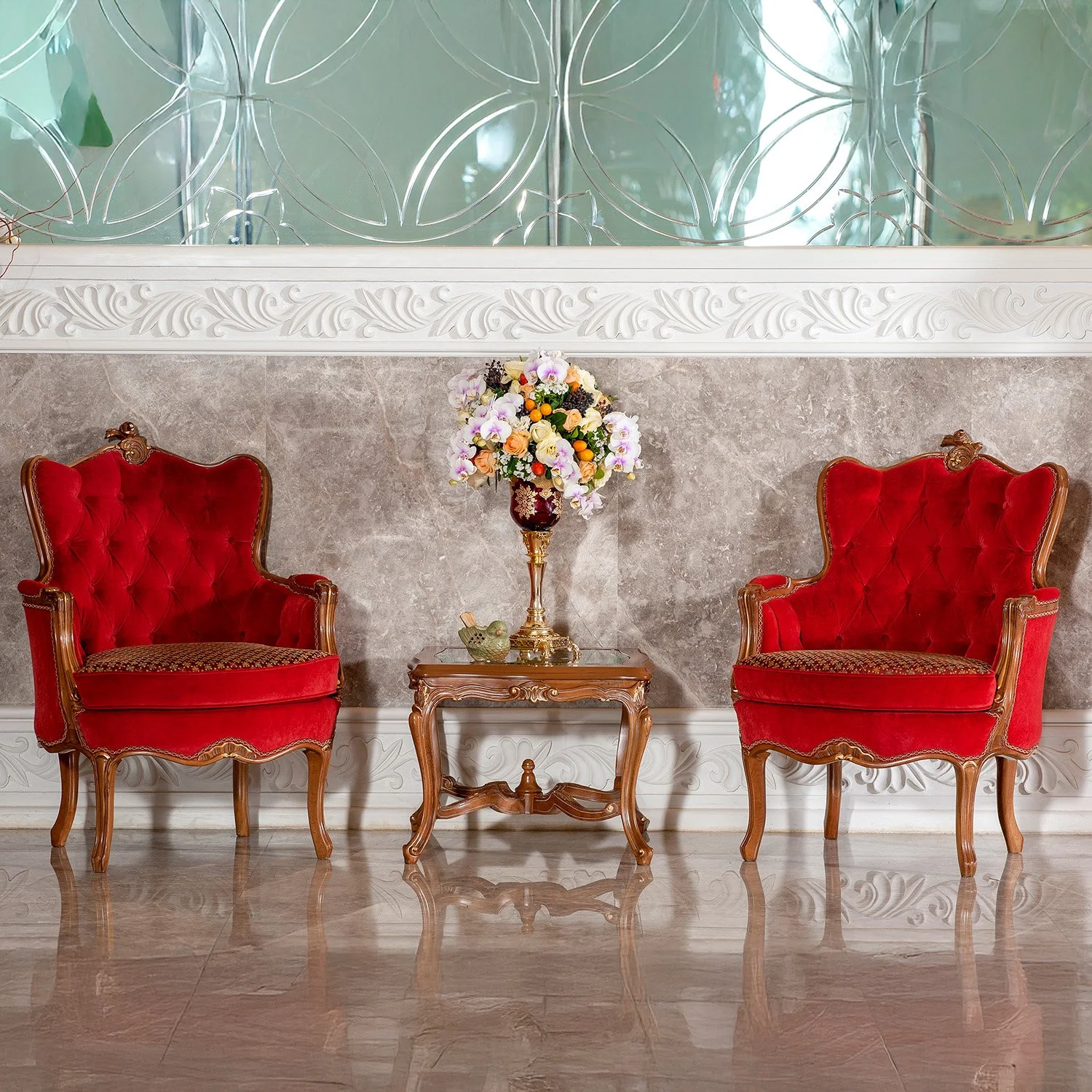 Two ornate, red velvet high-backed armchairs with carved wooden frames flank a small wooden side table with a colorful floral arrangement, set against a grey marble wall with decorative white molding and a reflective etched glass background.
