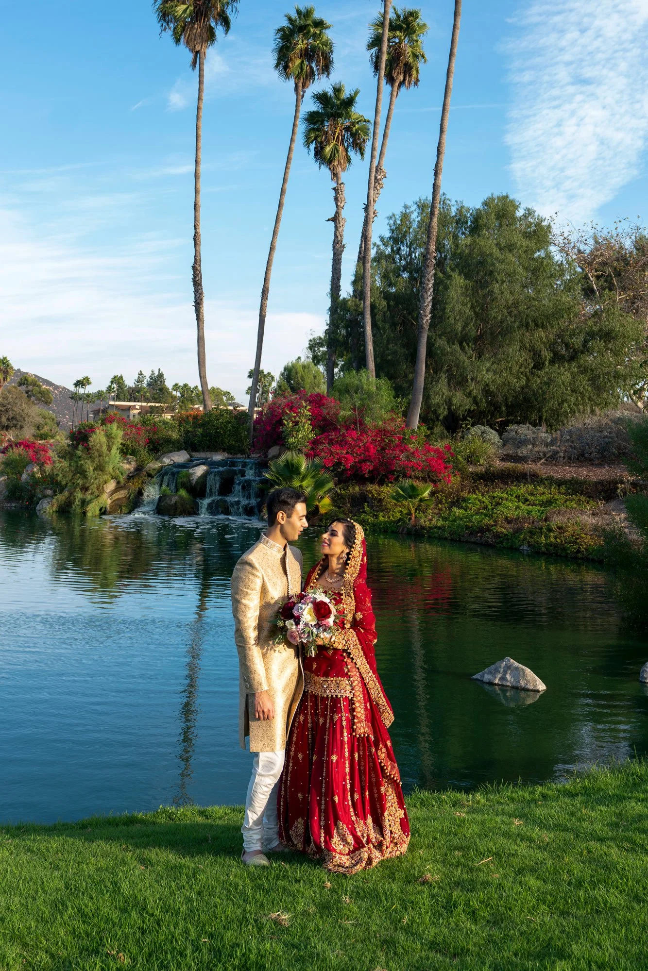A full-length photograph of a newly married couple posing outdoors in traditional attire. The groom wears a cream-colored outfit with gold accents, and the bride wears an ornate, deep red and gold lehenga. They stand on a grassy lawn beside a calm po