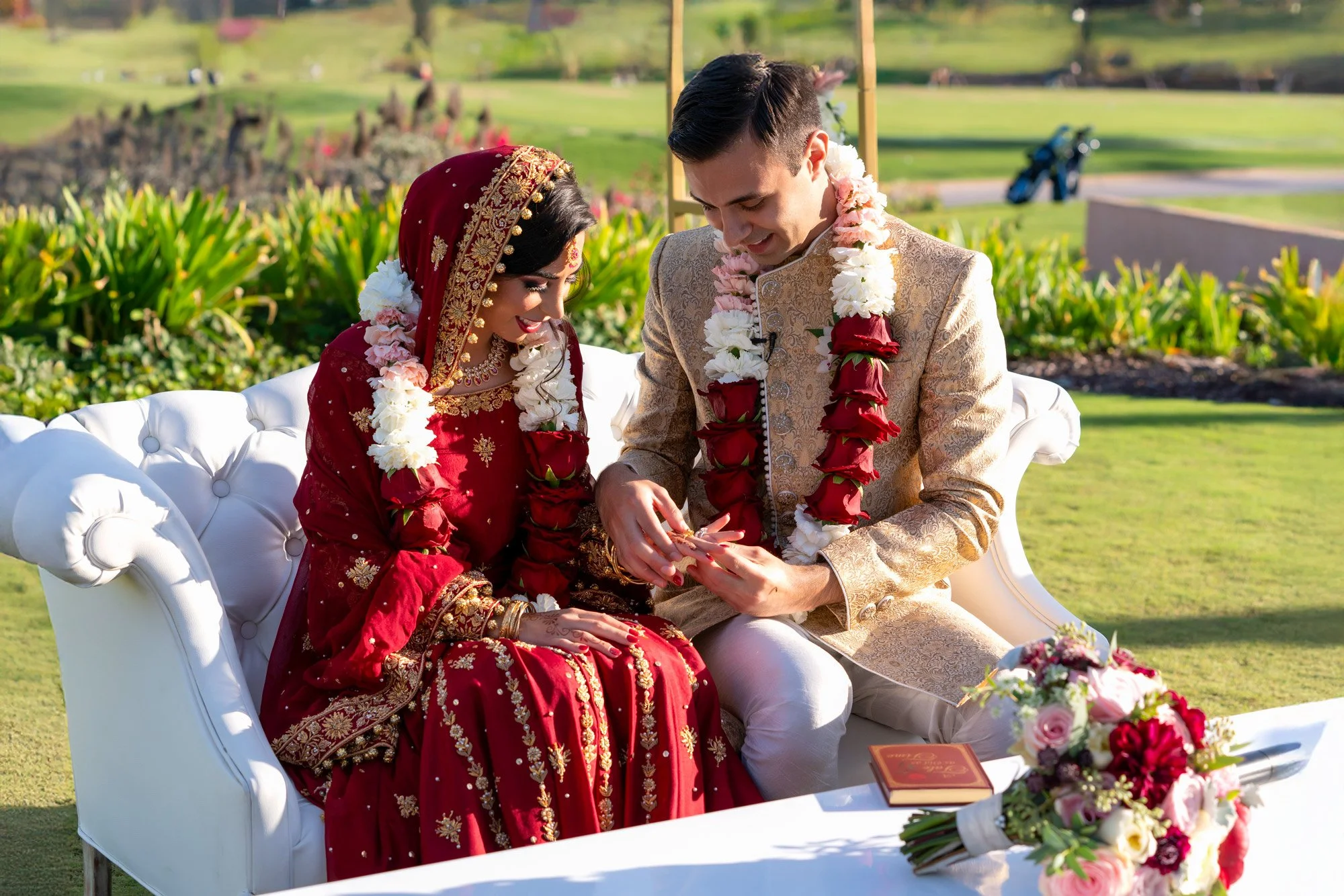 An outdoor photograph capturing the moment a groom places a ring on his bride's finger during a wedding ceremony. The couple is seated on a white tufted sofa. The groom wears a gold sherwani and the bride wears an ornate, deep red lehenga with gold e