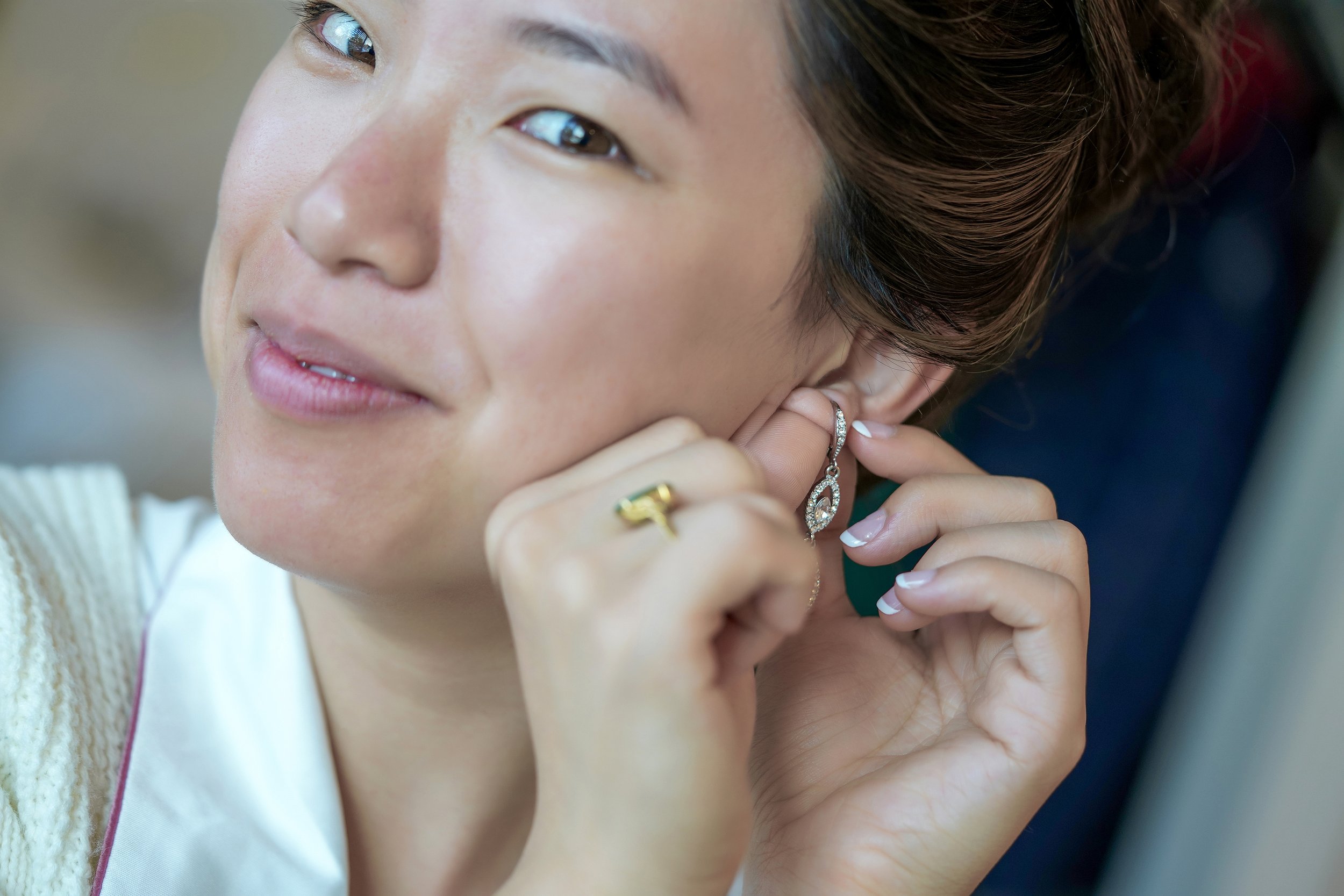 A close-up, slightly blurred photo of a smiling woman with her hair in an updo as she puts on a sparkling teardrop earring. Her hands are visible in the foreground, showing a gold ring on her finger and a neat French manicure. She is wearing a white 