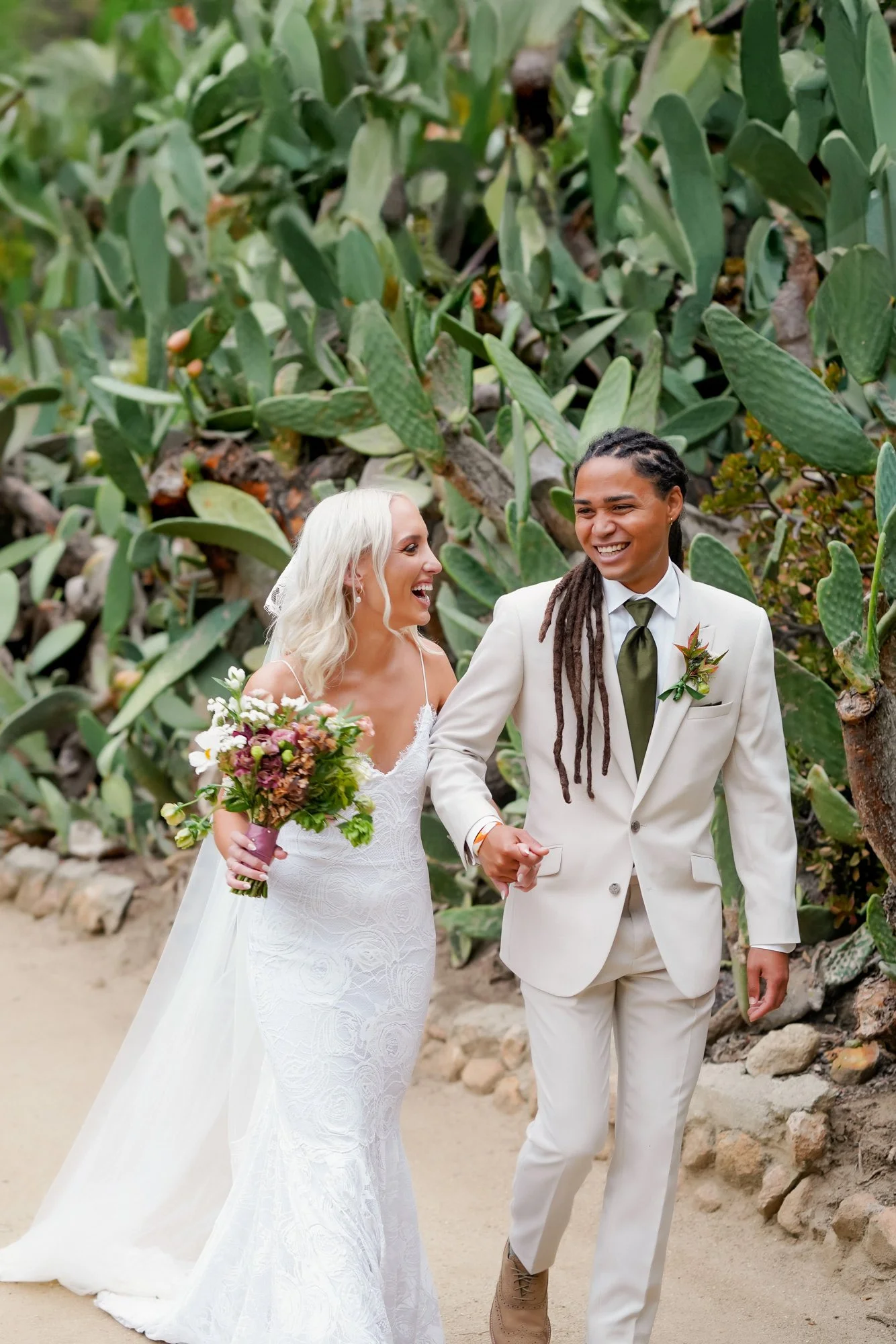 A photo of a newly married couple walking hand-in-hand down a dirt path, smiling and laughing together, surrounded by large prickly pear cacti. One person wears a white lace wedding dress with a veil and carries a bouquet, and the other wears a cream