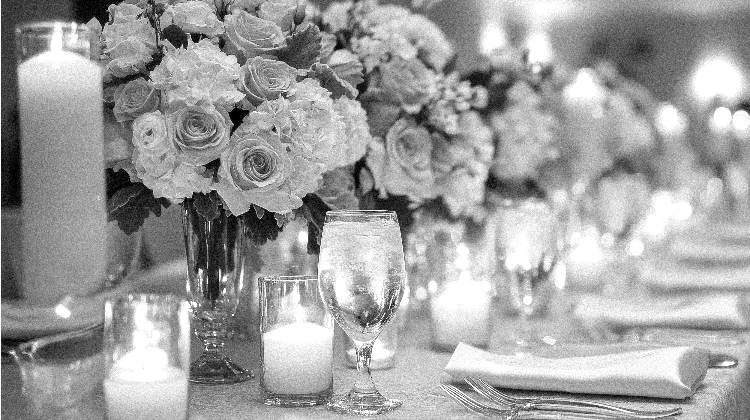 A black and white photograph of an elegant wedding table setting. The focus is on a floral centerpiece of roses and hydrangeas, surrounded by tall pillar candles and small votive candles. A water glass, napkin, and cutlery are visible at a formally s