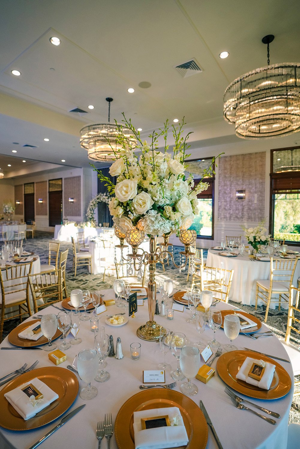 An elegantly set dining table for a formal event. The table features gold charger plates, white napkins, gold-rimmed glasses, and a prominent gold candelabra centerpiece with white floral arrangements. Other set tables, large windows, and ornate circ