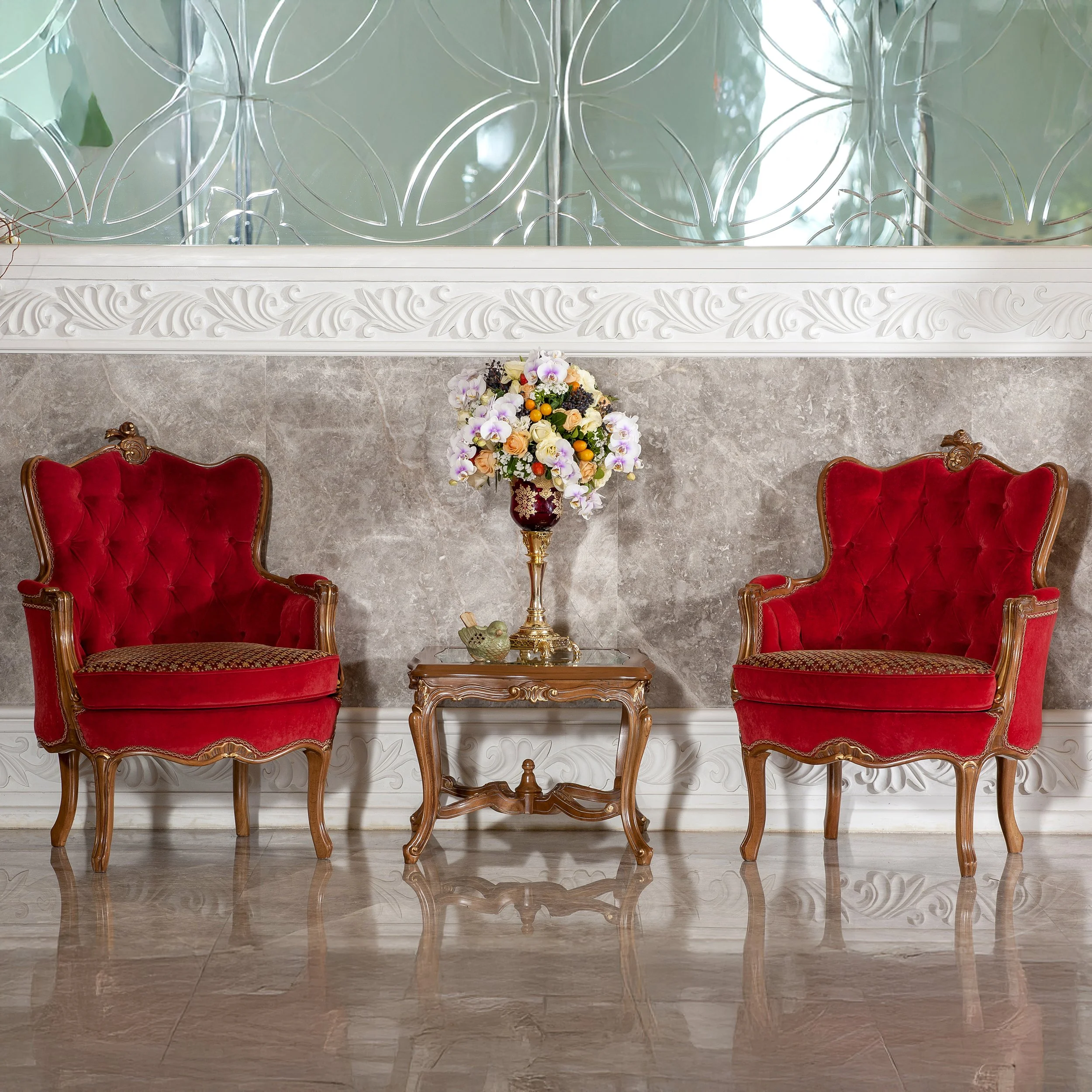 Two ornate, red velvet high-backed armchairs with carved wooden frames flank a small wooden side table with a colorful floral arrangement, set against a grey marble wall with decorative white molding and a reflective etched glass background.
