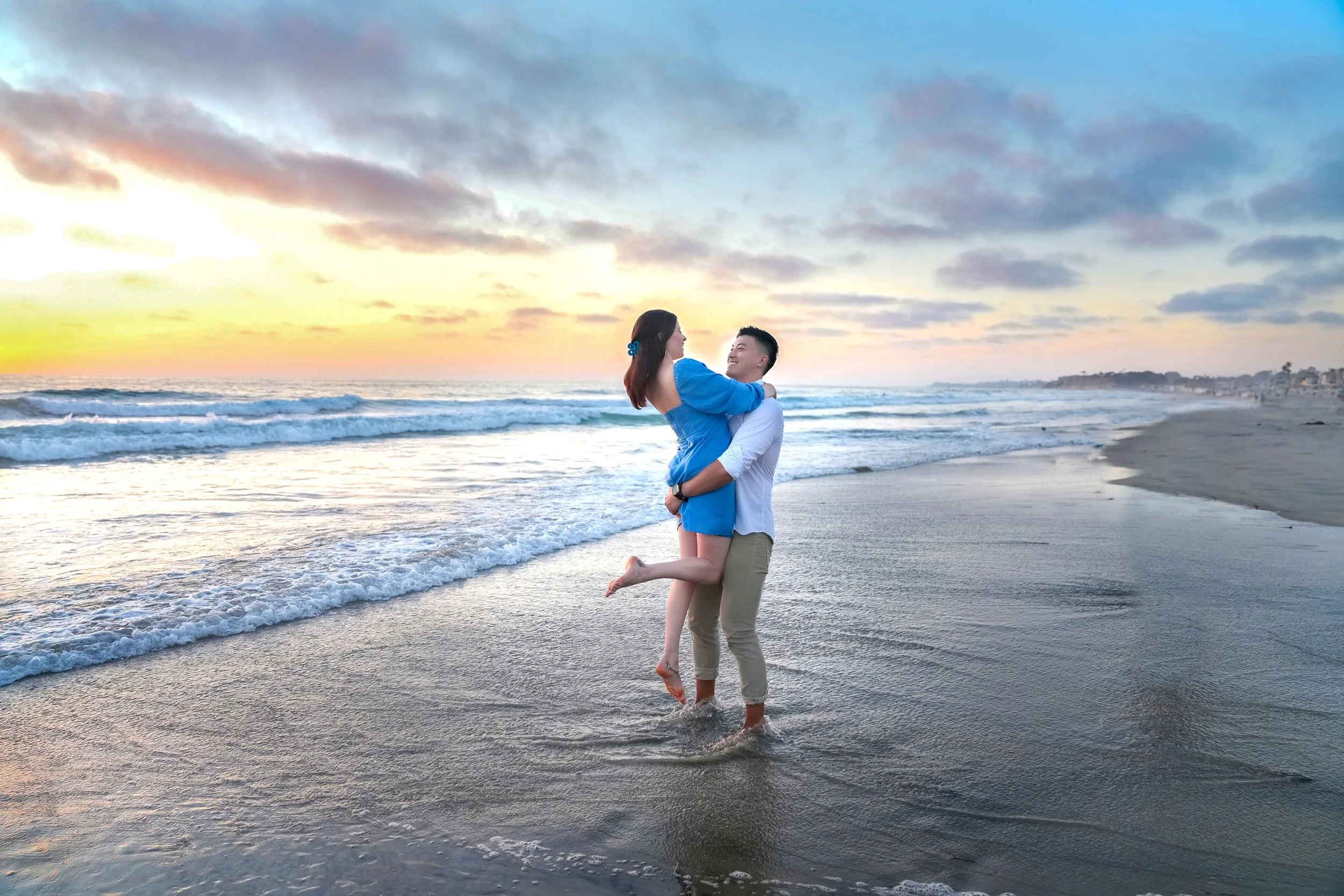 A man lifts and hugs a woman in a blue dress while standing in the shallow ocean water at Del Mar beach during a vibrant sunset engagement session.