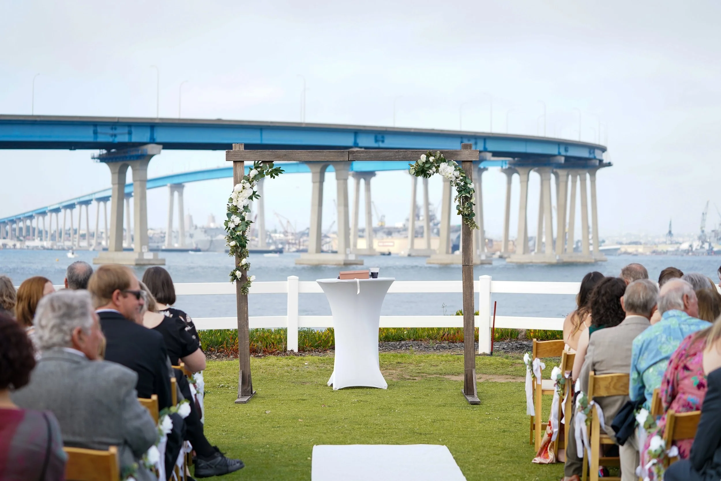 An outdoor wedding ceremony setup on a green lawn with a wooden arch decorated with white flowers, white guest chairs, and the large blue San Diego-Coronado Bridge spanning the water in the background.