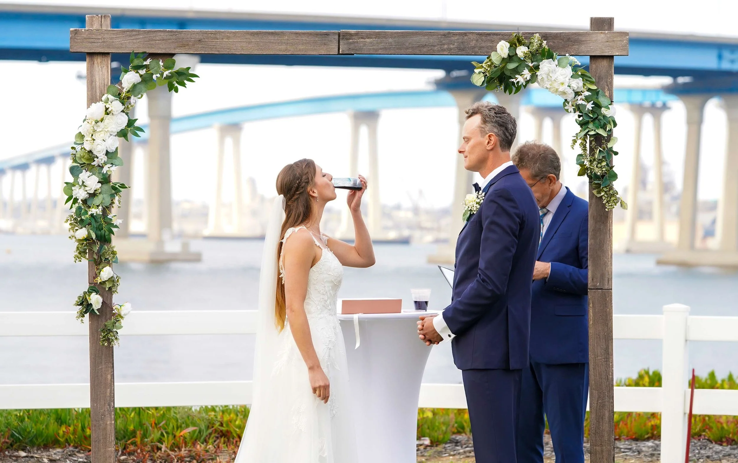 A bride in a white wedding gown takes a drink from a dark bottle during an outdoor ceremony, standing with the groom and officiant under a wooden arch with Coronado Bridge visible in the background over the water.


