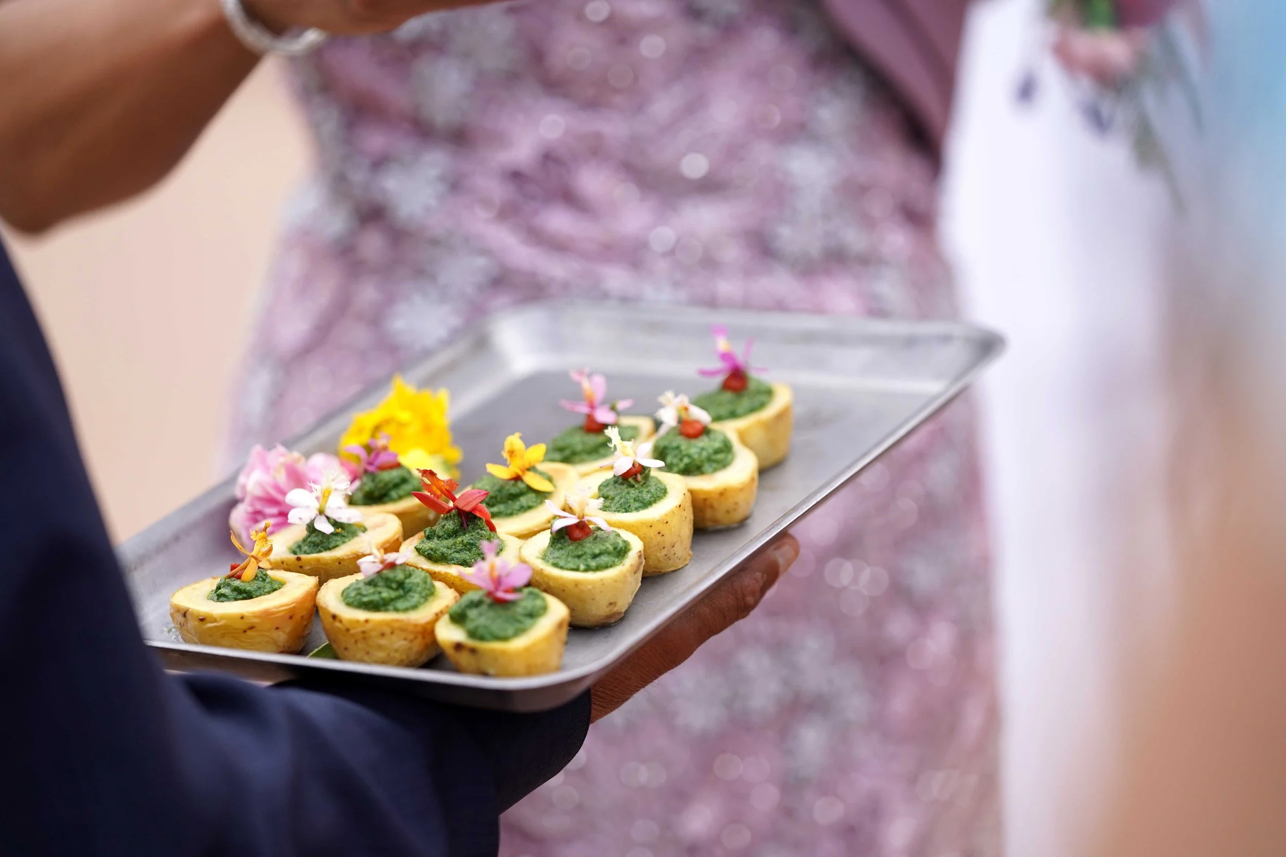 A metal tray held by a person in a dark suit contains several mini, bite-sized appetizers, each topped with a green sauce and small edible flowers.