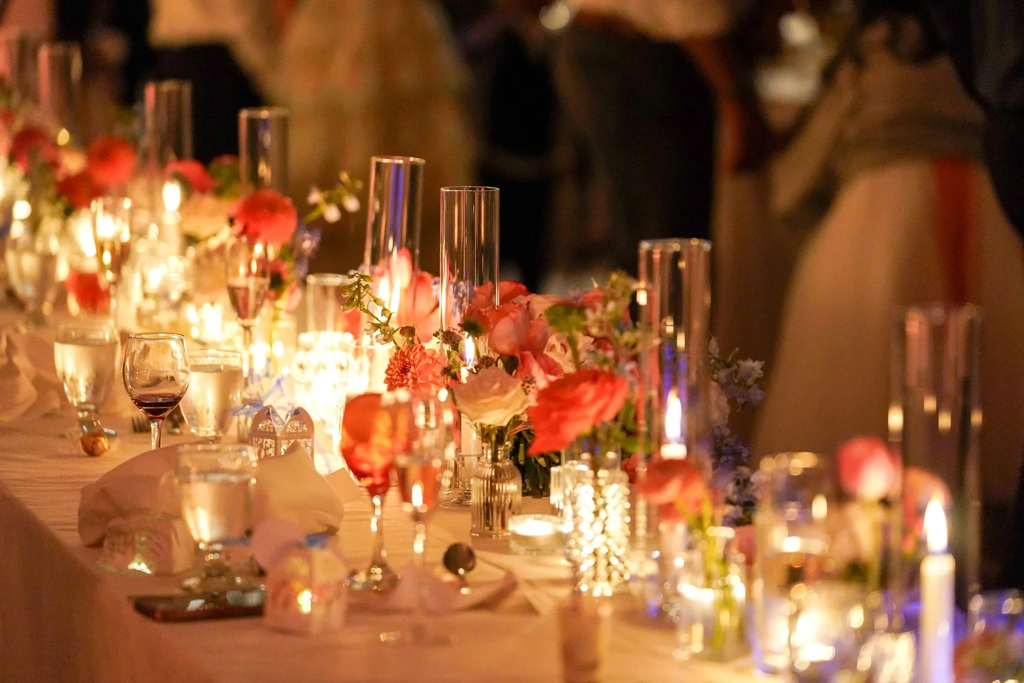 A long, formal dining table set for an evening event with a white tablecloth, featuring warm candlelight from numerous glass holders, arrangements of pink, red, and white flowers, glassware for drinks, and blurred figures of guests in the background.