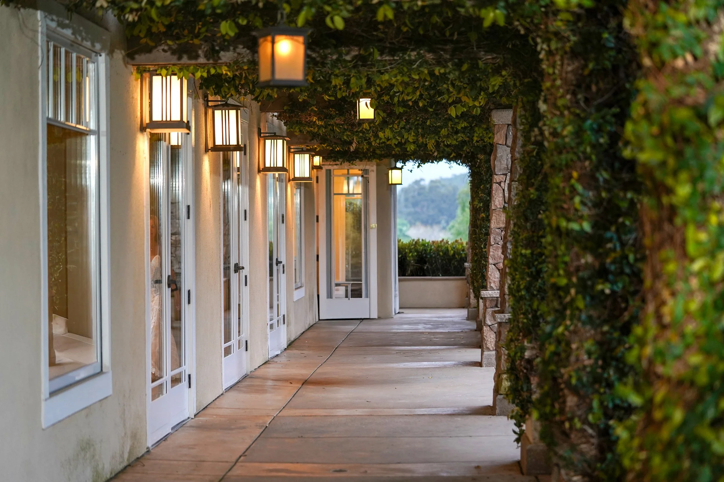 A covered outdoor walkway or veranda with a stucco wall on the left and a stone column wall on the right, featuring several illuminated, rustic-style lanterns hanging from an ivy-covered trellis ceiling at twin peak golf course.
