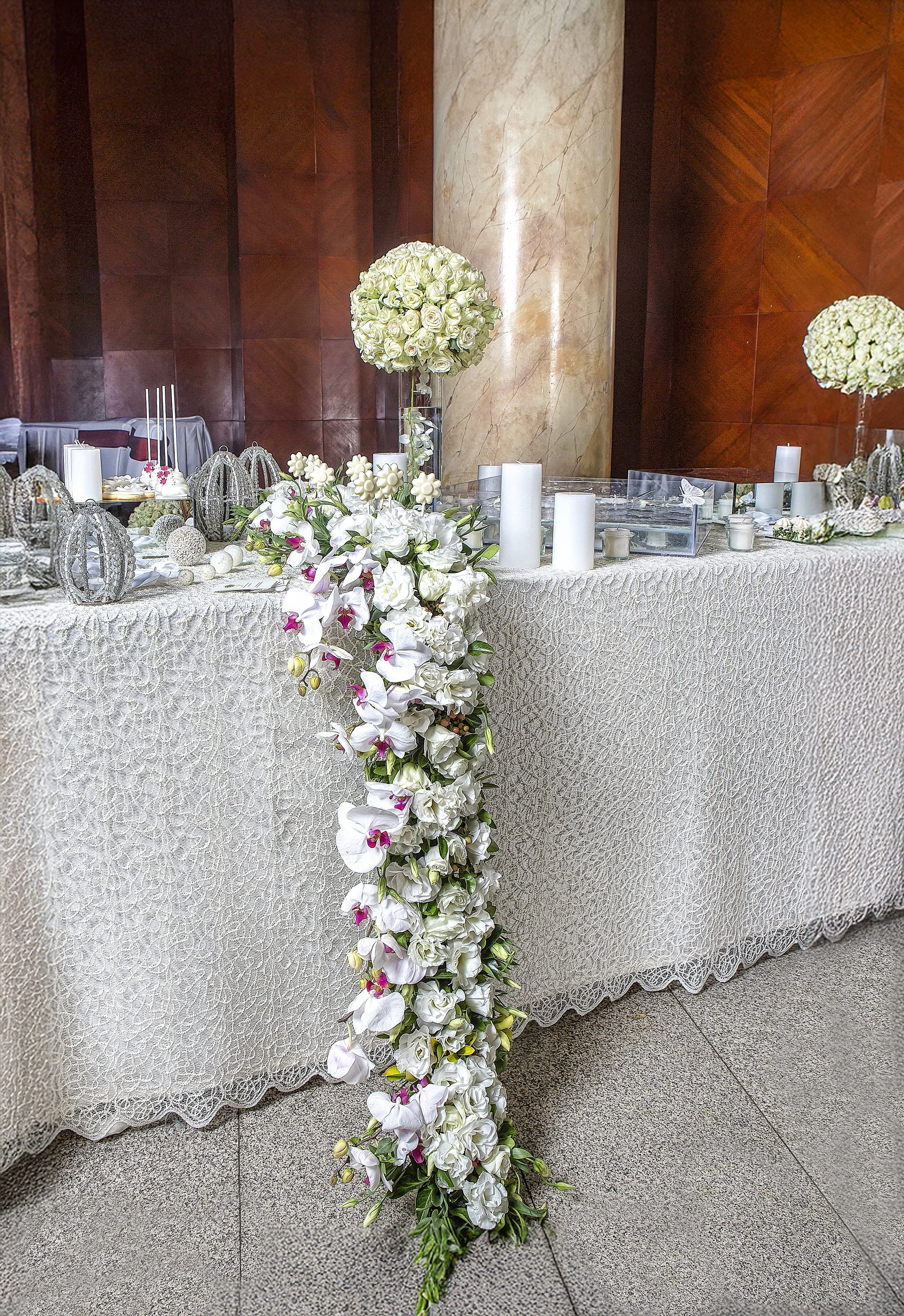 A formal event table draped in a white lace tablecloth, featuring a dramatic cascade of white roses and orchids with magenta accents, alongside white pillar candles, silver sphere decorations, and a background of dark wood paneling and a marble colum