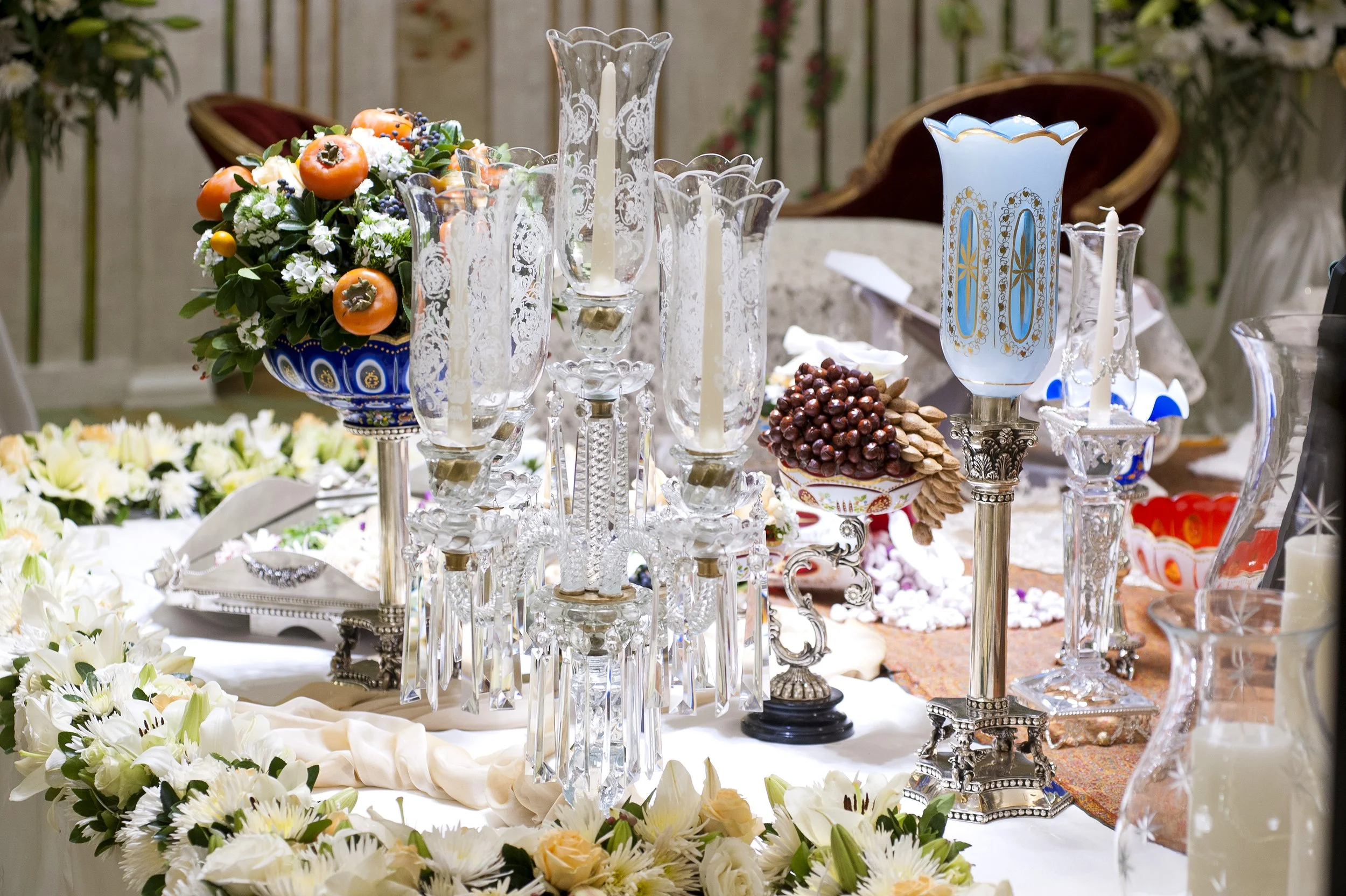An ornate Persian wedding table setting (Sofreh Aghd) featuring crystal candelabras, a blue and gold candle holder, a bowl of nuts and dried fruits, and extensive white and orange floral arrangements.