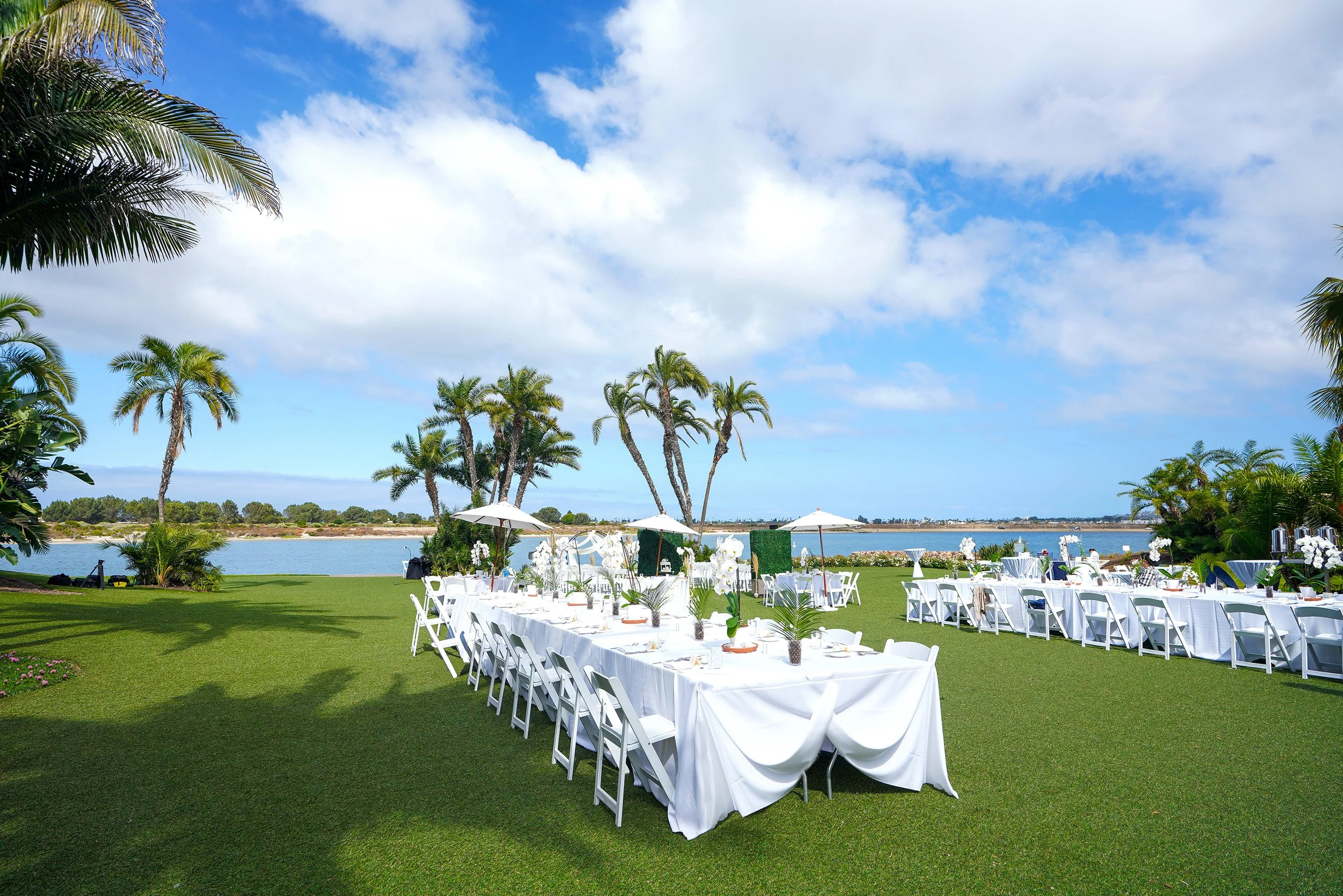 An outdoor wedding reception is set up on a lush green lawn overlooking a calm body of water under a bright, partly cloudy sky. Long dining tables draped in elegant white linens are decorated with tropical floral arrangements and surrounded by white 