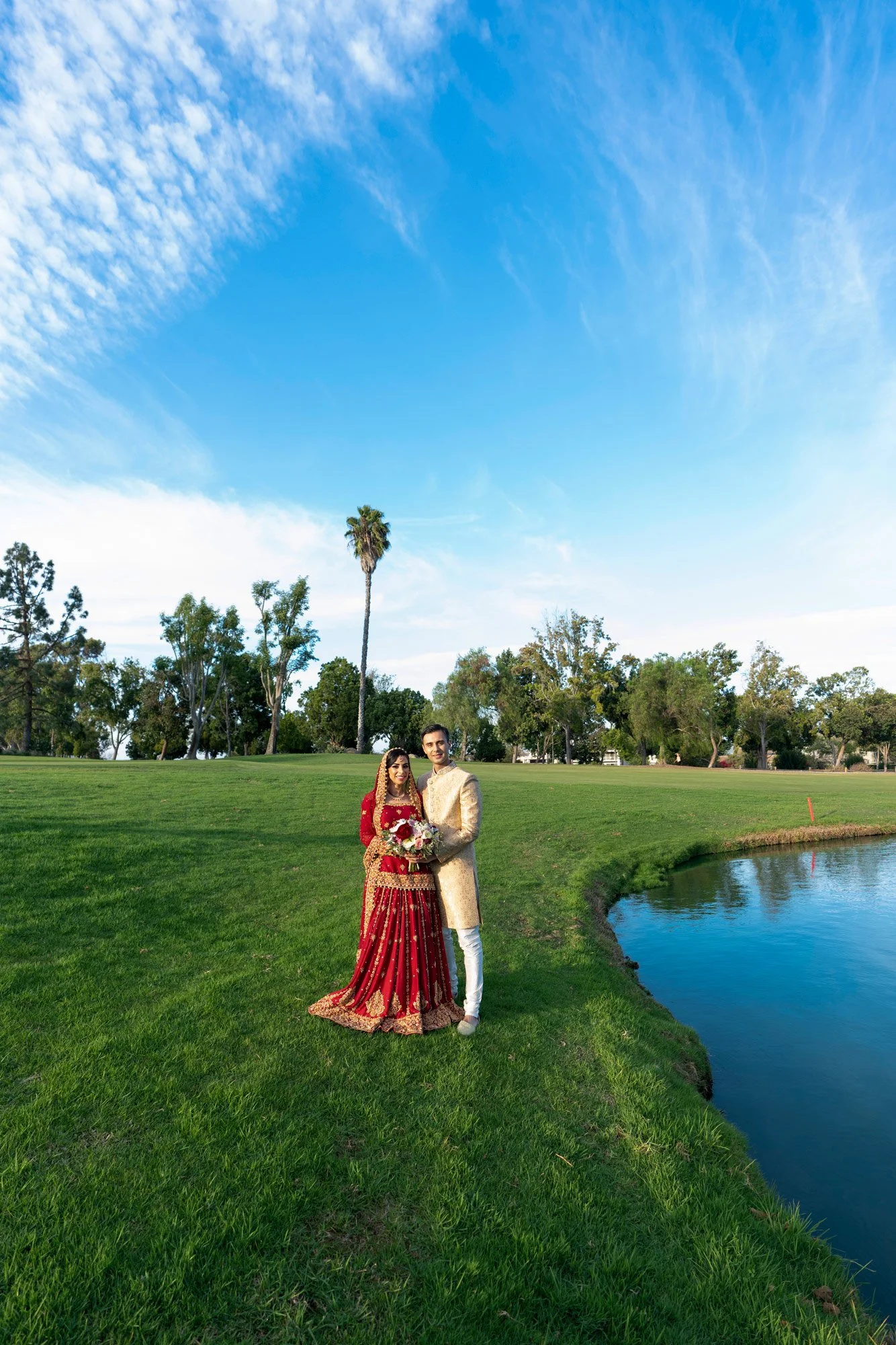 A full-length photograph of a newly married couple posing outdoors in traditional attire. The groom wears a cream-colored outfit with gold accents, and the bride wears an ornate, deep red and gold lehenga. They stand on a grassy lawn beside a calm po