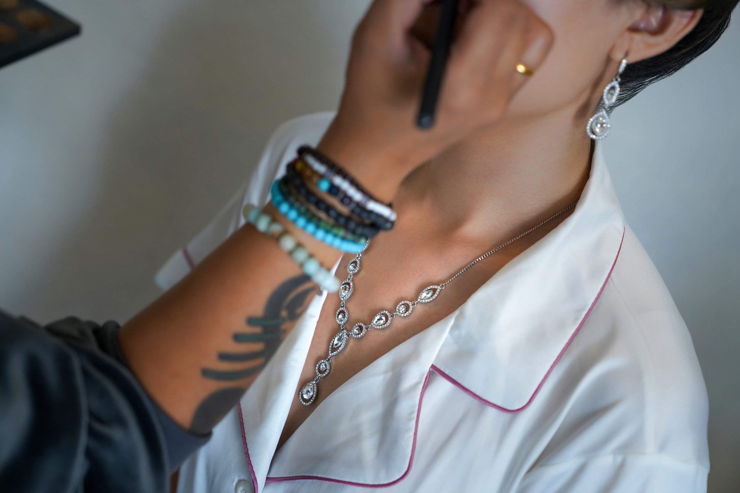 A close-up shot of a person in white pajamas with pink piping receiving a makeup application. A hand with a tribal-style tattoo and multiple beaded bracelets is visible in the foreground, holding a makeup brush to the person's face. The person being 