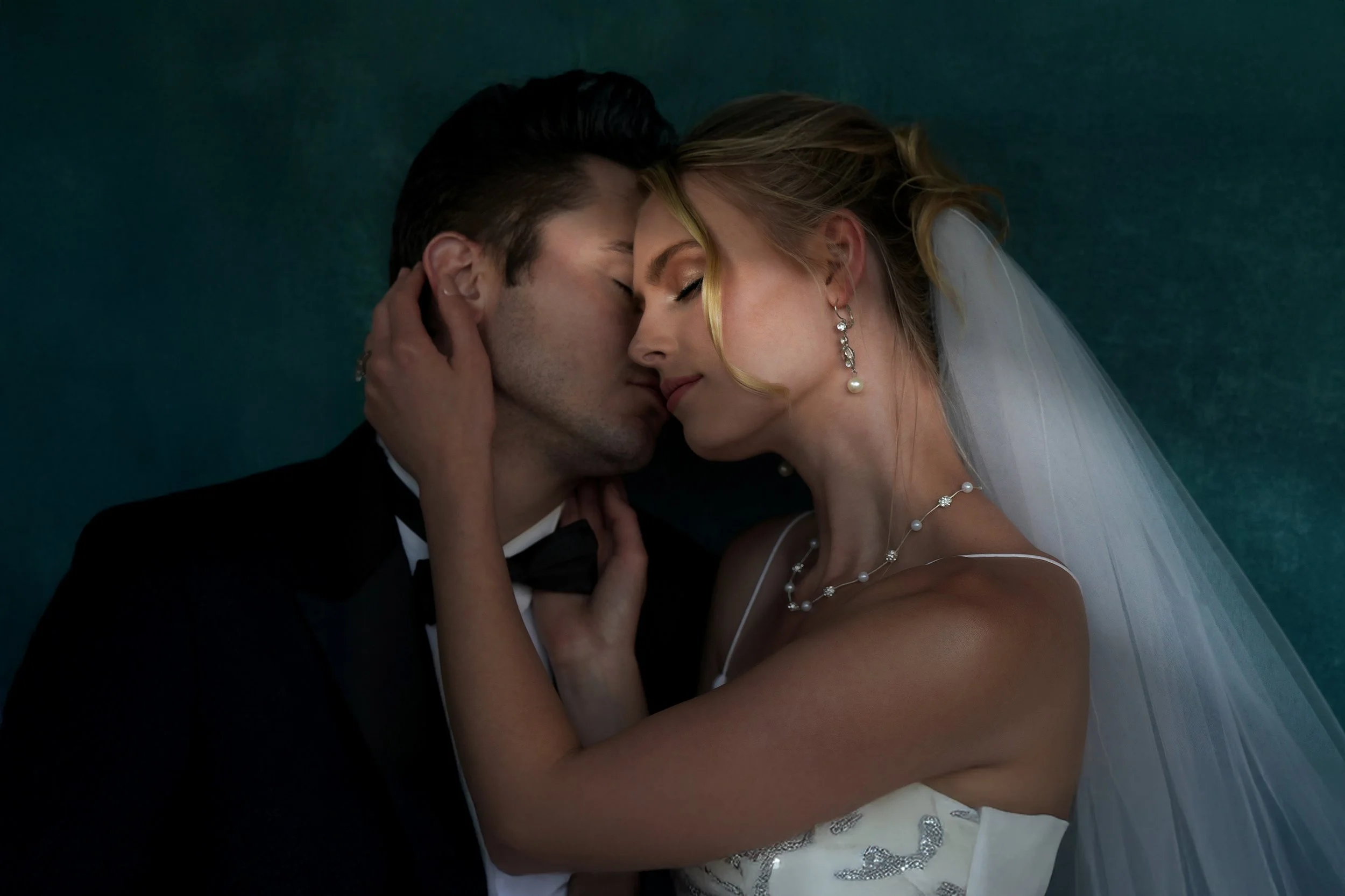 A bride in a white wedding dress and groom in a black tuxedo stand closely together with their foreheads touching.