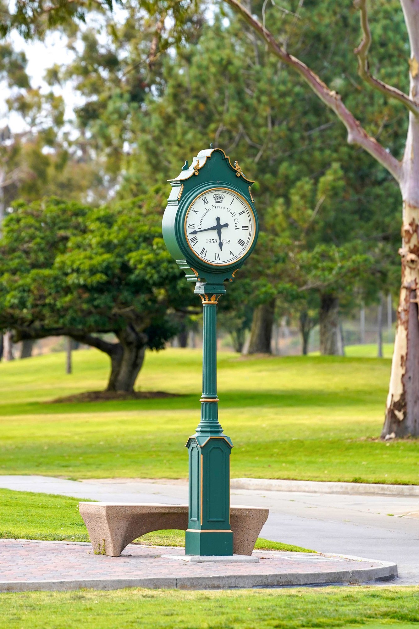 A tall, vintage-style street clock painted emerald green with gold trim sits on a paved walkway next to a concrete bench in a Coronado golf course setting.