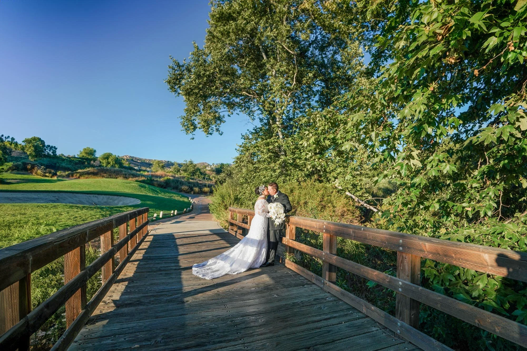 A full-length outdoor photograph of a newly married couple kissing on a wooden footbridge on a sunny day. The bride wears a white lace wedding dress with long sleeves and a flowing train and holds a white floral bouquet, while the groom wears a black