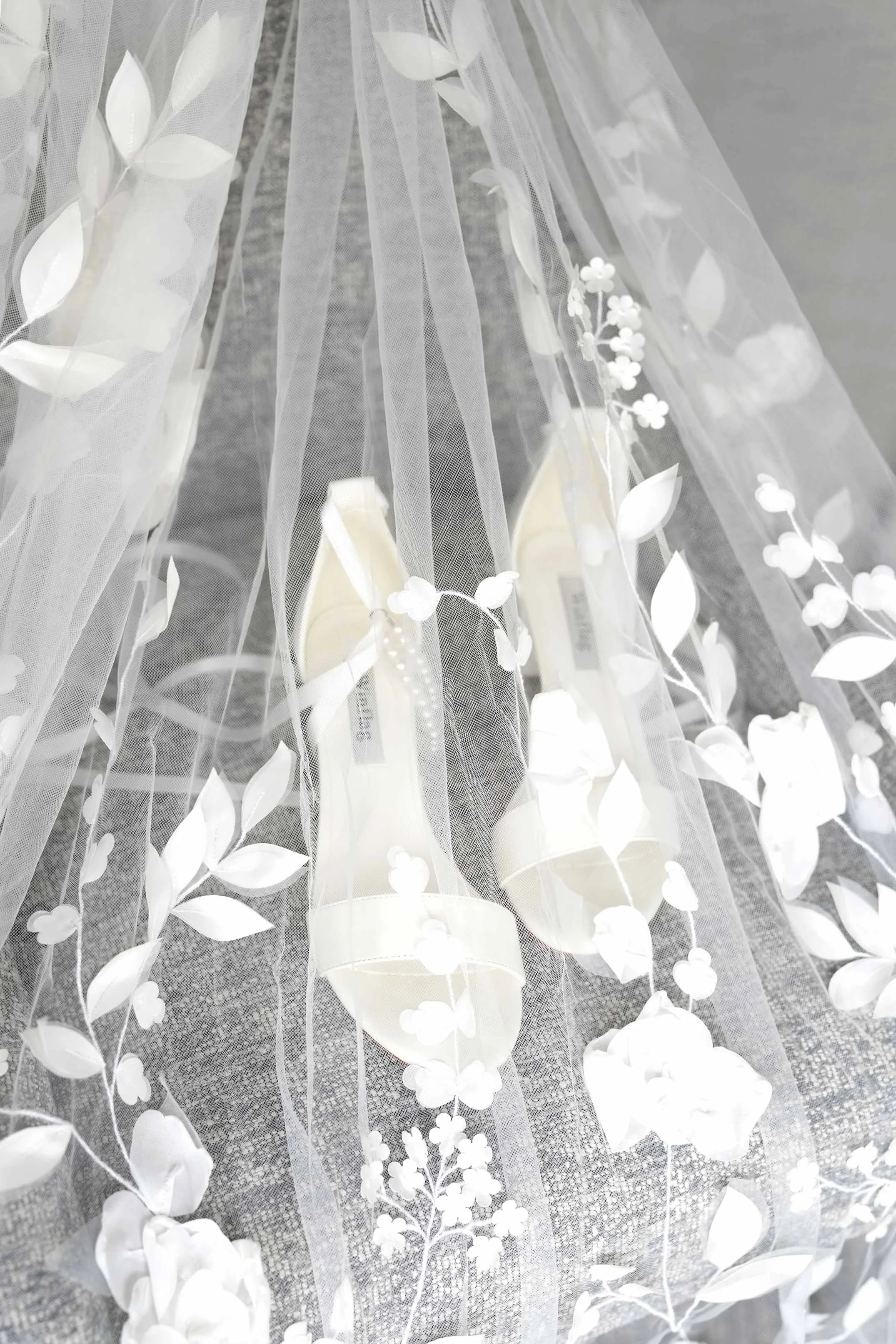 A pair of white high-heeled sandals featuring floral details and an ankle strap sits beneath a white wedding veil with large lace flower appliques.