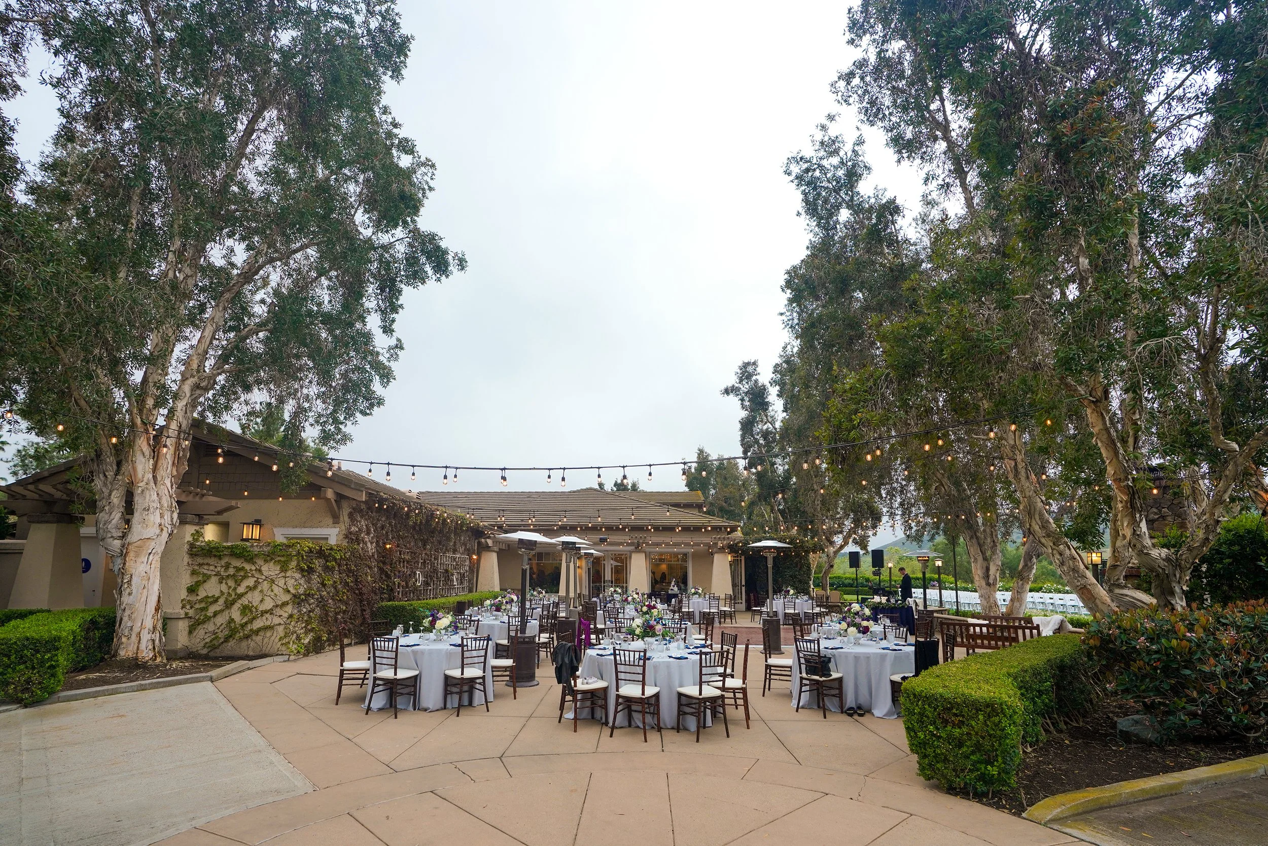 Numerous round tables are arranged across the patio, each covered with light grey or white tablecloths and paired with dark wooden event chairs at twin oaks golf course.