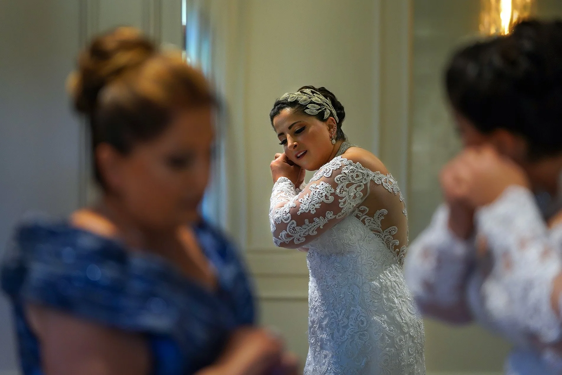A bride in an intricate lace off-the-shoulder gown puts on her earrings while preparing for her wedding. She wears a large, sparkling silver leaf-shaped headpiece in her dark hair, which is styled in an updo. The foreground shows two other people sof