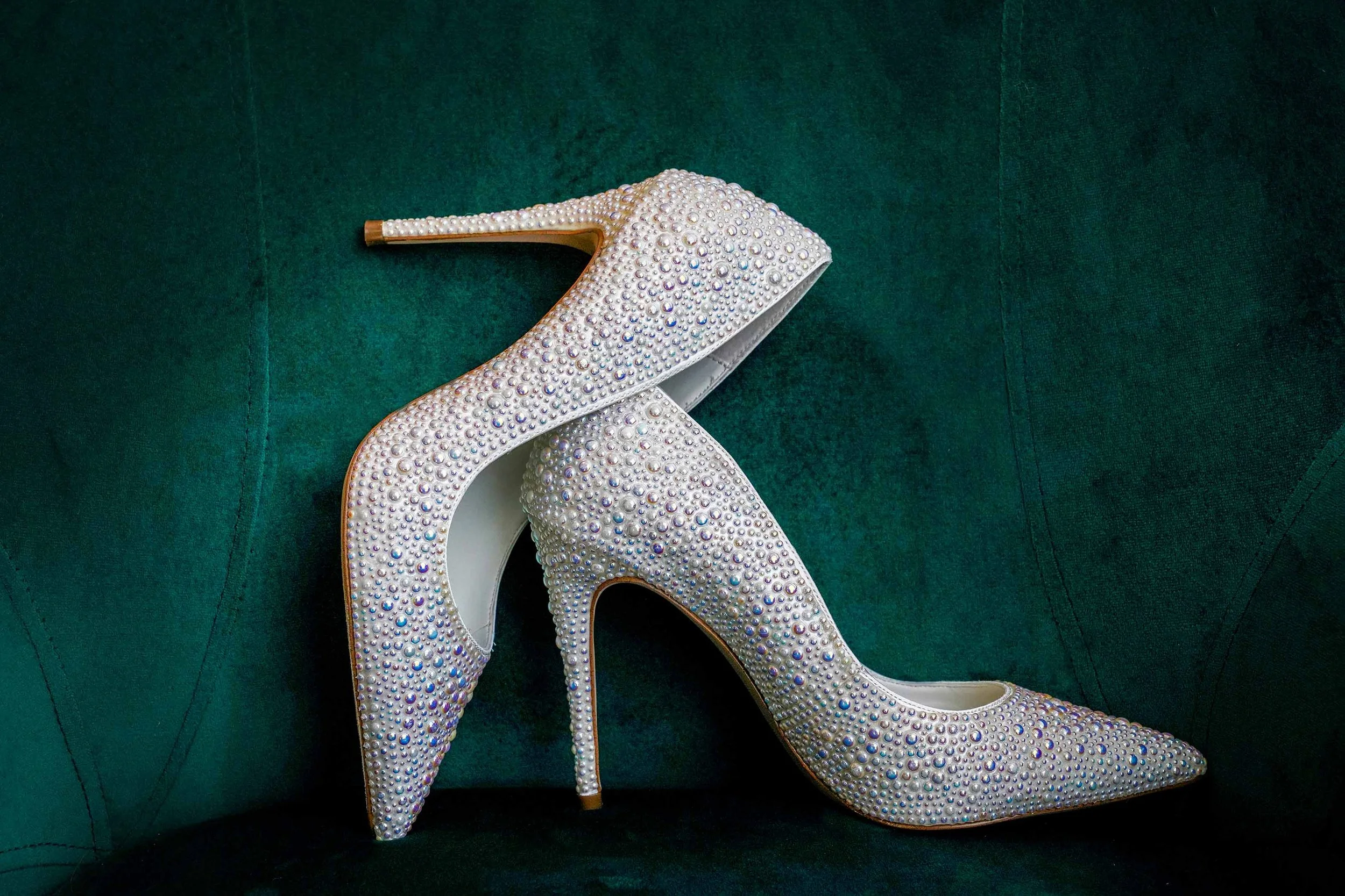 A close-up photograph of a pair of pointed-toe stiletto pumps, completely covered in iridescent white and colorful rhinestones and pearls, set against a dark emerald-green velvet background.
Betsey Johnson | Shoes | Blue By Betsey Johnson Shine Rhine