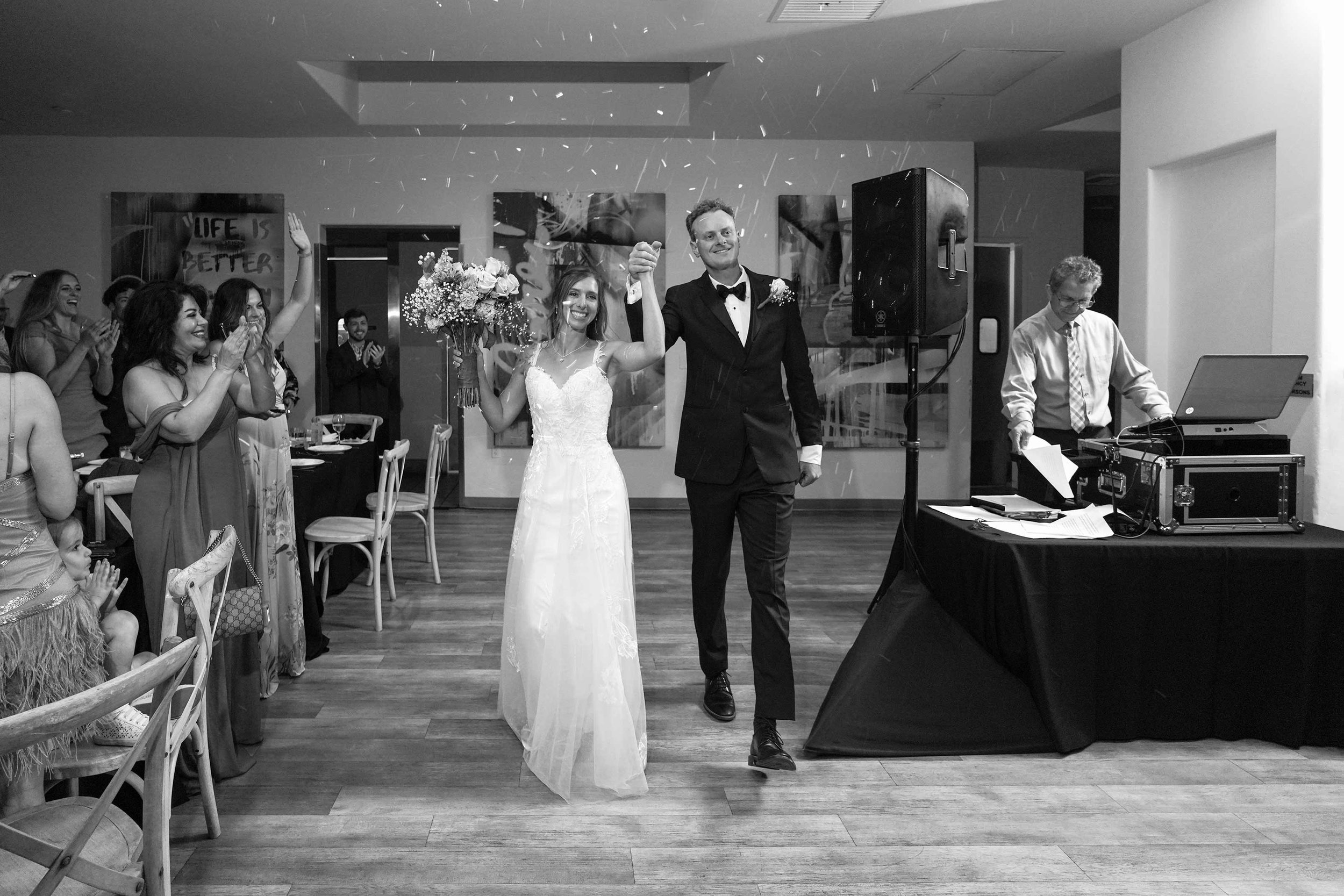 A black and white photograph of a bride in a white lace dress and a groom in a tuxedo walking down a hallway holding hands, smiling, while confetti falls and guests applaud.


