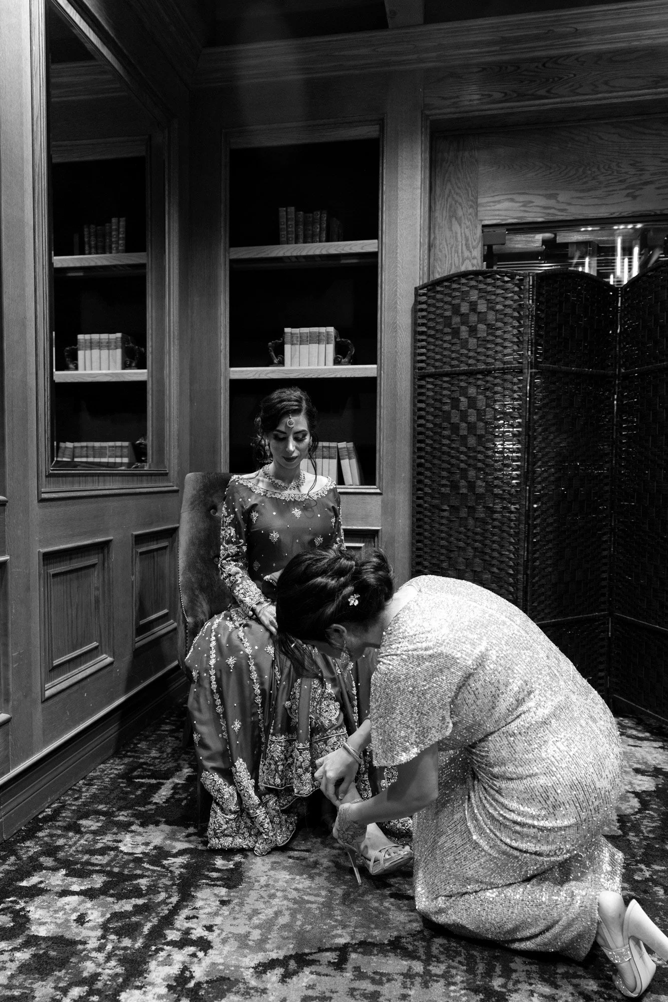 A black and white photograph in a wood-paneled room with bookshelves, where a person is kneeling down to help a seated person with their shoes. The seated person wears a dark, ornate dress, and the kneeling person wears a shimmering dress. A room div