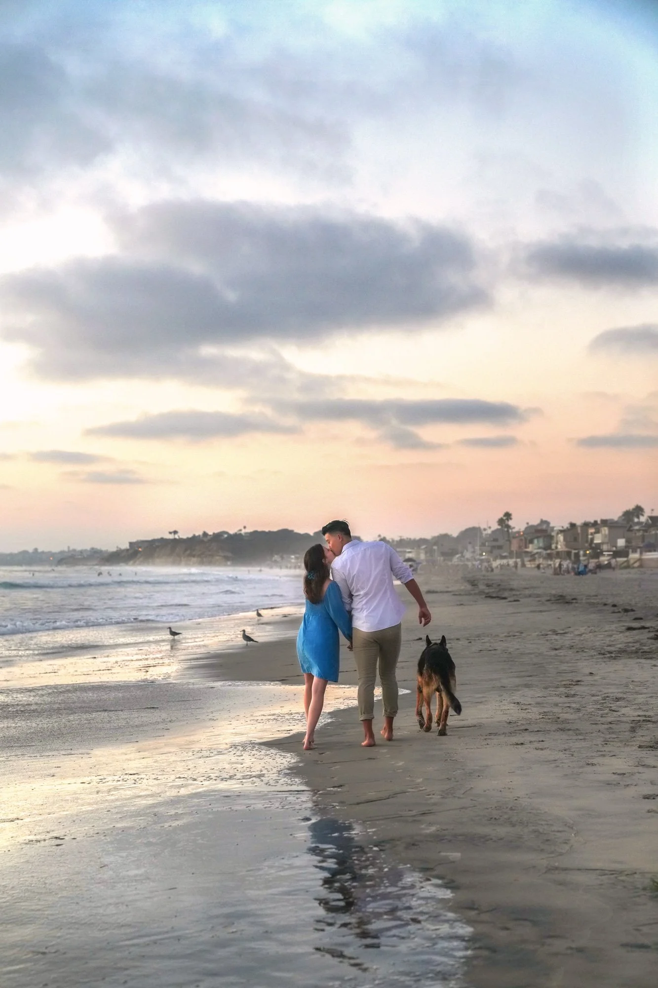 An outdoor photograph of a couple and their dog walking on a sandy beach at sunset or sunrise. The woman wears a blue dress, the man wears a white shirt and tan pants, and a dark dog walks beside them. They are holding hands and kissing. The image ca