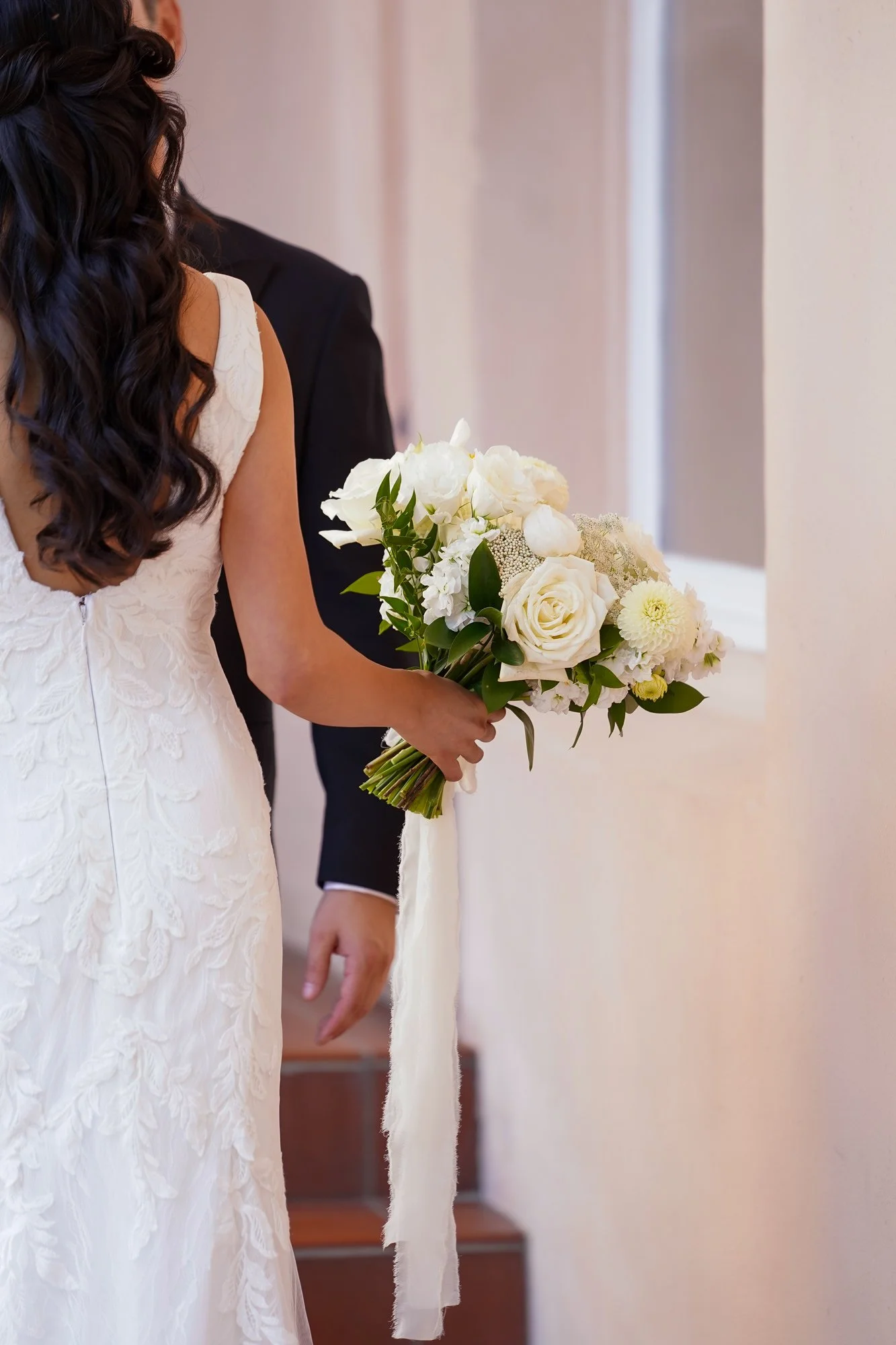 A photograph taken from behind a bride in a white lace wedding dress as she holds a white floral bouquet. She is walking alongside a man in a black suit down a set of steps. The bouquet is tied with a long white ribbon at Hotel La Valencia, La Jolla.