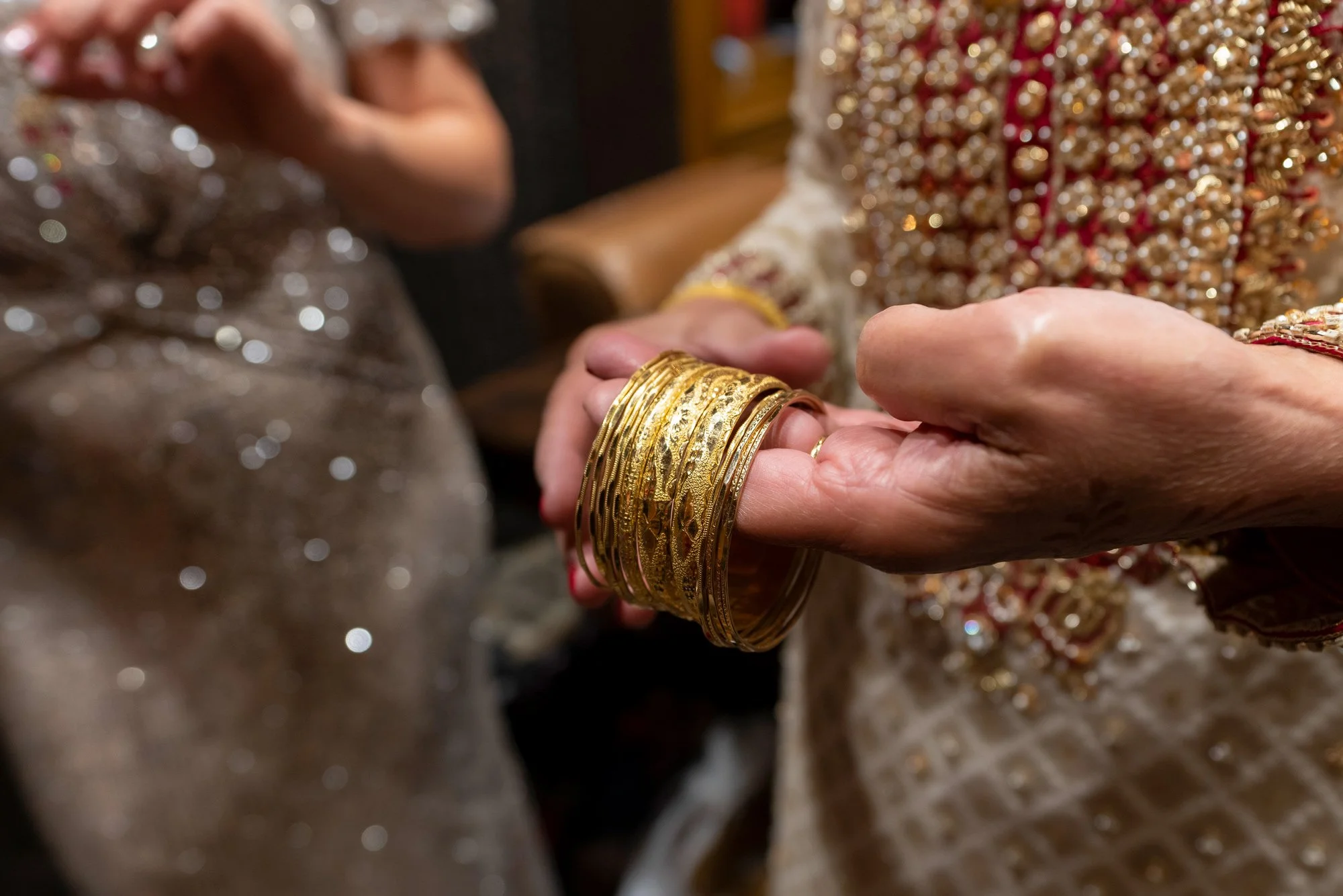 A close-up photograph of a hand holding a stack of gold bangles. A person in an ornate, cream-colored, and red outfit is holding the bangles, appearing to place them on or take them off their wrist. Another person in a light, sequined dress is visibl