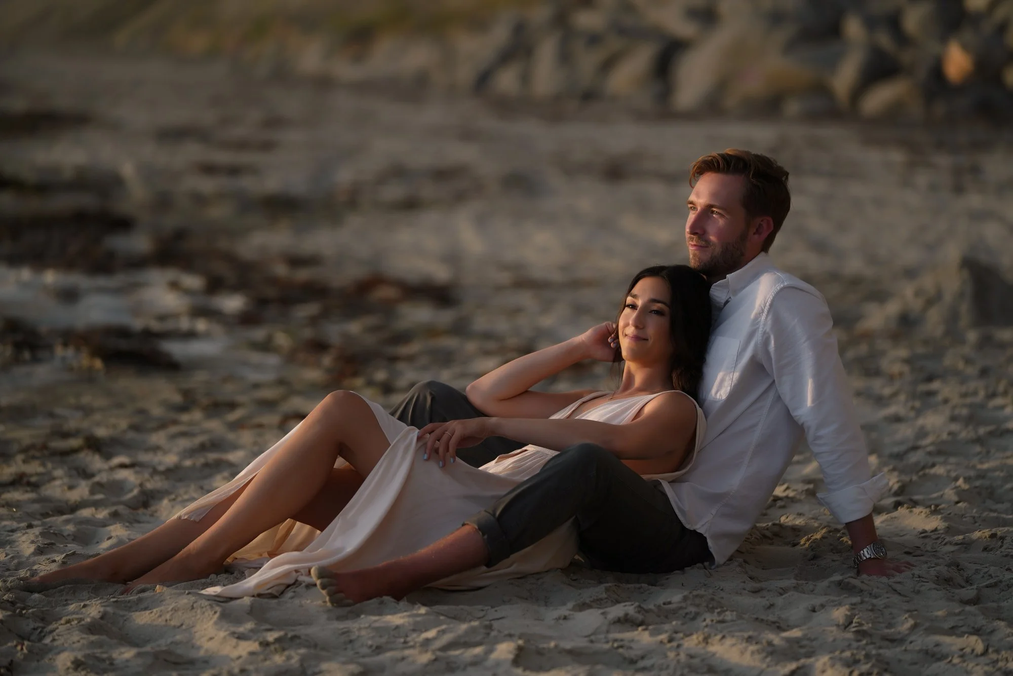 An outdoor photograph of a couple sitting on a sandy beach during sunset or sunrise. The woman, in a light-colored dress, leans against the man, who wears a white long-sleeved shirt and dark pants and is looking out towards the light. They are sittin
