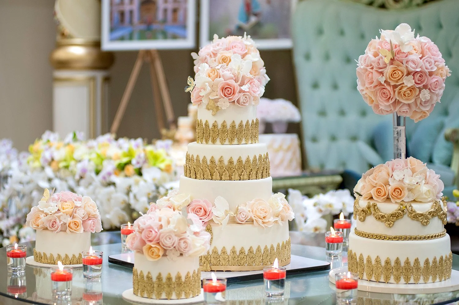 A five-tiered white and gold wedding cake and several smaller matching single-tiered cakes, all decorated with pink and white roses, were displayed on a glass table surrounded by red votive candles.
