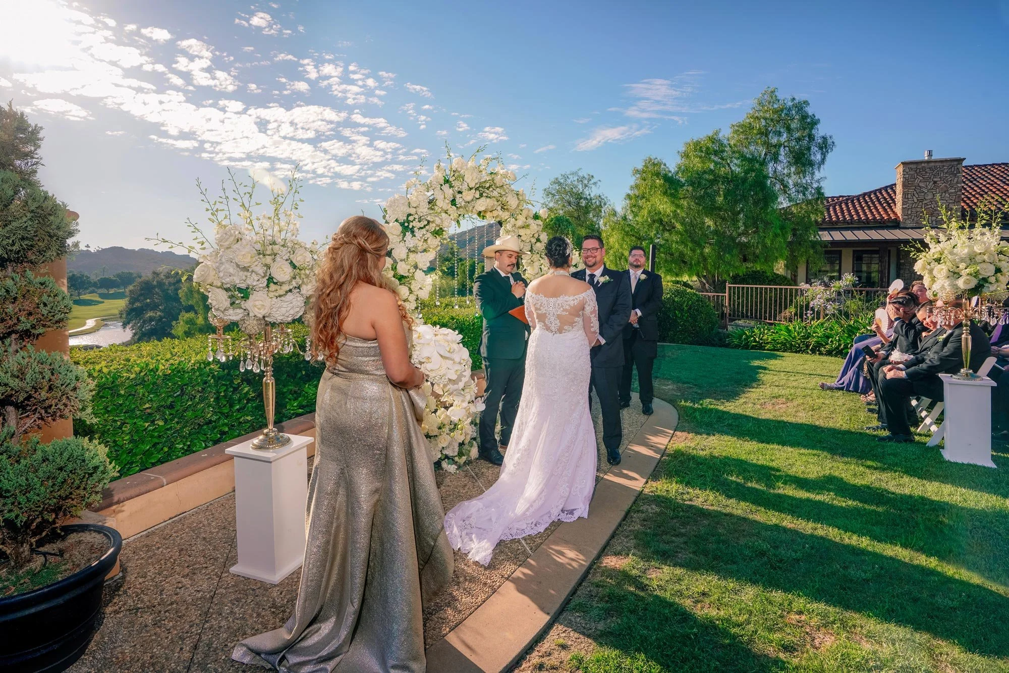 An outdoor photograph capturing a wedding ceremony on a sunny day. The bride, in a white lace dress, and groom, in a black suit, stand at the altar area facing an officiant. A woman in a silver dress stands in the foreground, facing away from the cam