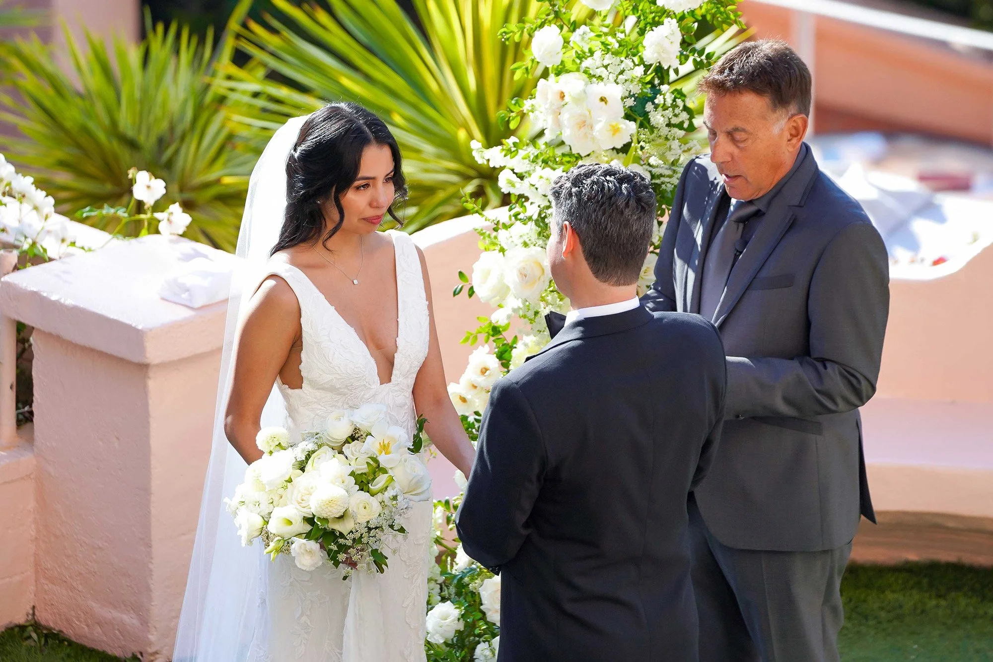 An outdoor photograph of a bride in a white lace wedding dress, holding a white floral bouquet, facing the groom during a ceremony. A man in a gray suit stands beside the groom, likely the officiant or a witness. The scene is framed by white floral a