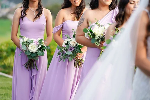 A line of bridesmaids wearing mismatched lavender dresses—including one-shoulder, off-the-shoulder, and spaghetti strap styles—hold bouquets of white and purple roses with green eucalyptus.
