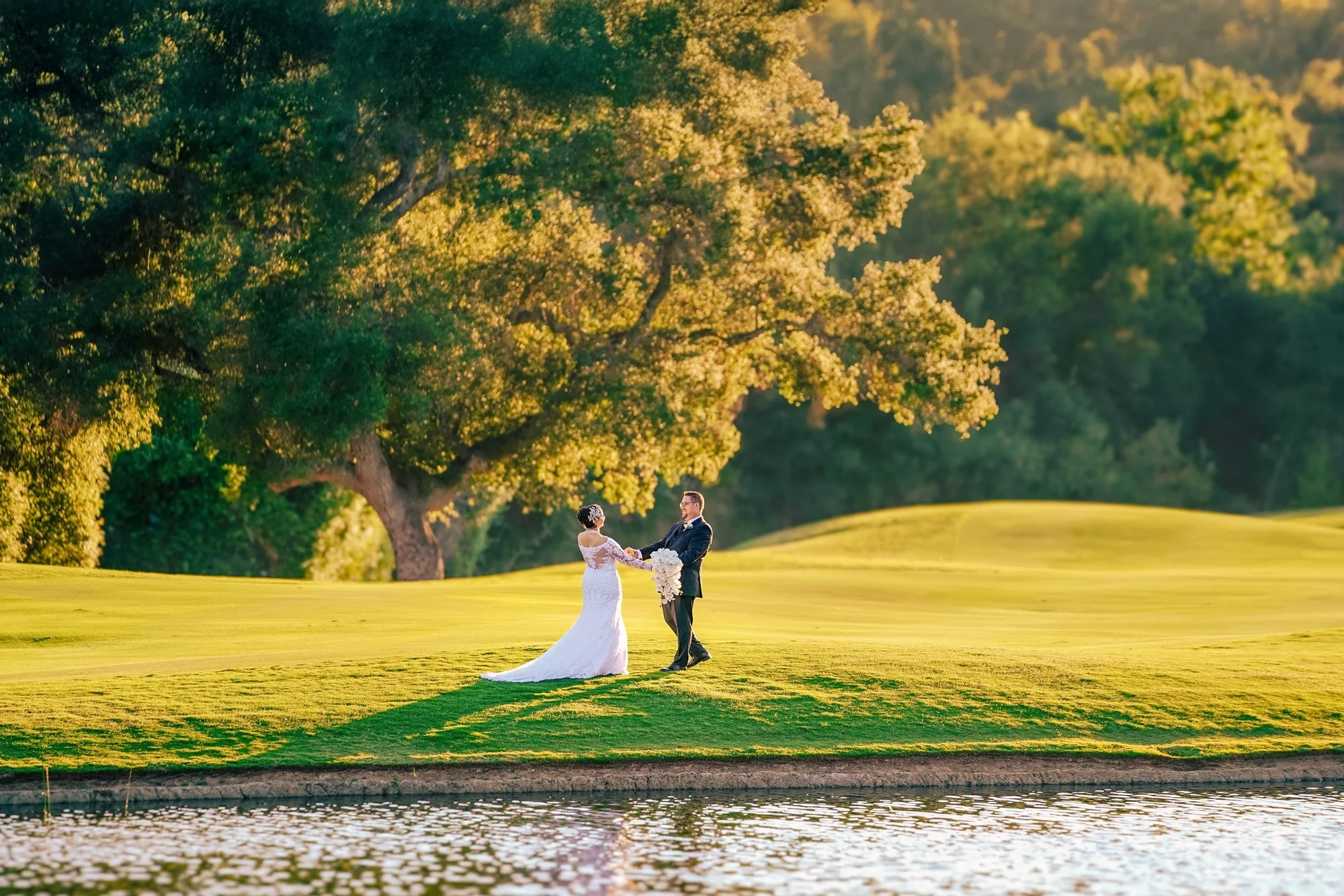 A full-length outdoor photograph of a newly married couple holding hands and looking at each other on a grassy knoll beside a body of water. The bride wears a white wedding dress with a long train and holds a white floral bouquet, while the groom wea
