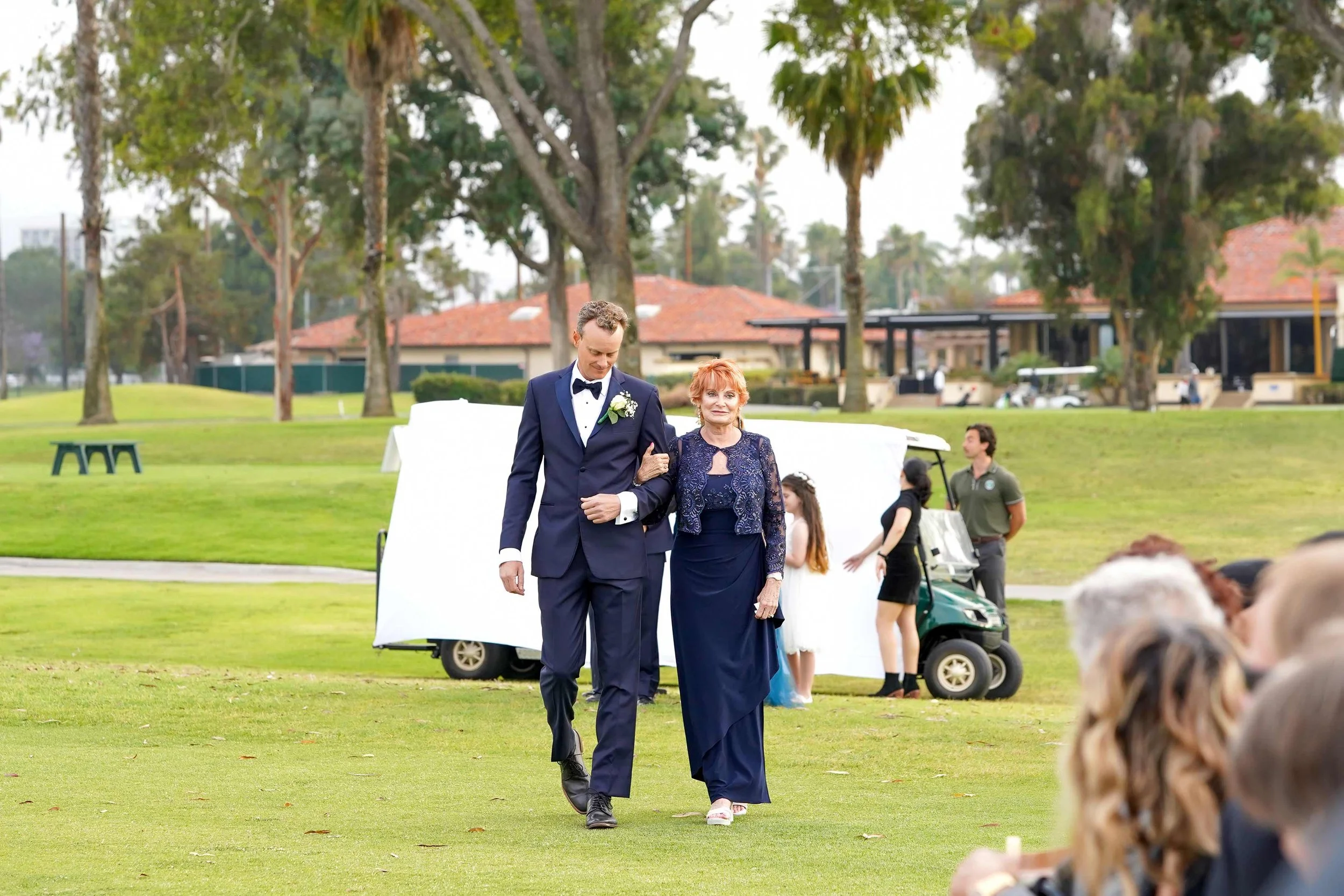The groom in a navy blue tuxedo and his mother in a matching navy blue dress and lace jacket walk arm-in-arm across a green lawn in Coronado Golf Club during the wedding procession.


