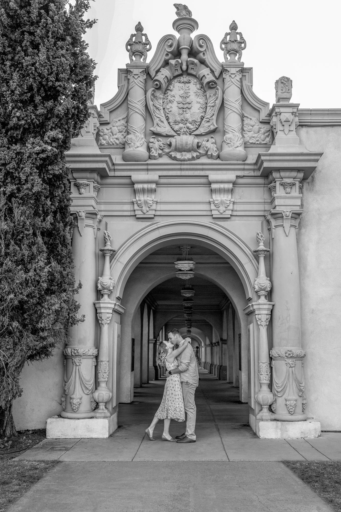 A black and white photograph of a man and woman embracing and kissing in an arched hallway with Spanish Colonial Revival architecture. The hallway features ornate stone pillars and arches, typical of those found in Balboa Park in San Diego. A dark tr