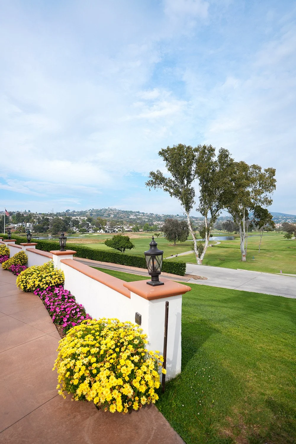 A scenic view from a patio overlooking a green golf course and rolling hills with houses, featuring vibrant yellow and pink flower beds and decorative black lanterns.