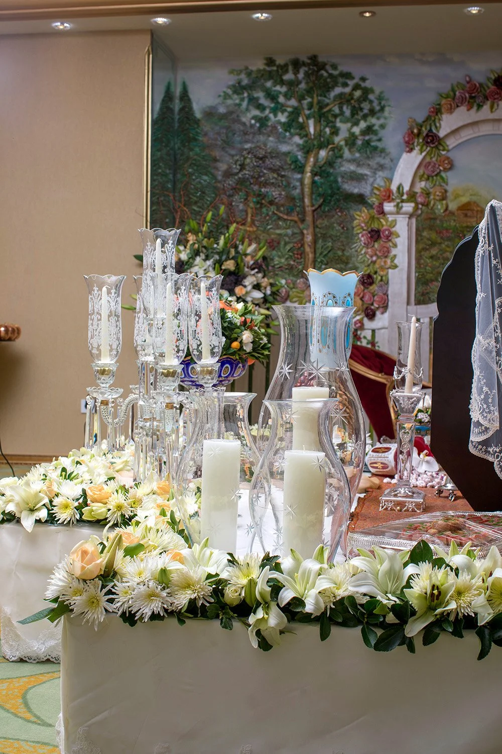 An ornate Persian wedding table setting (Sofreh Aghd) featuring crystal candelabras, a blue and gold candle holder, a bowl of nuts and dried fruits, and extensive white and orange floral arrangements.
