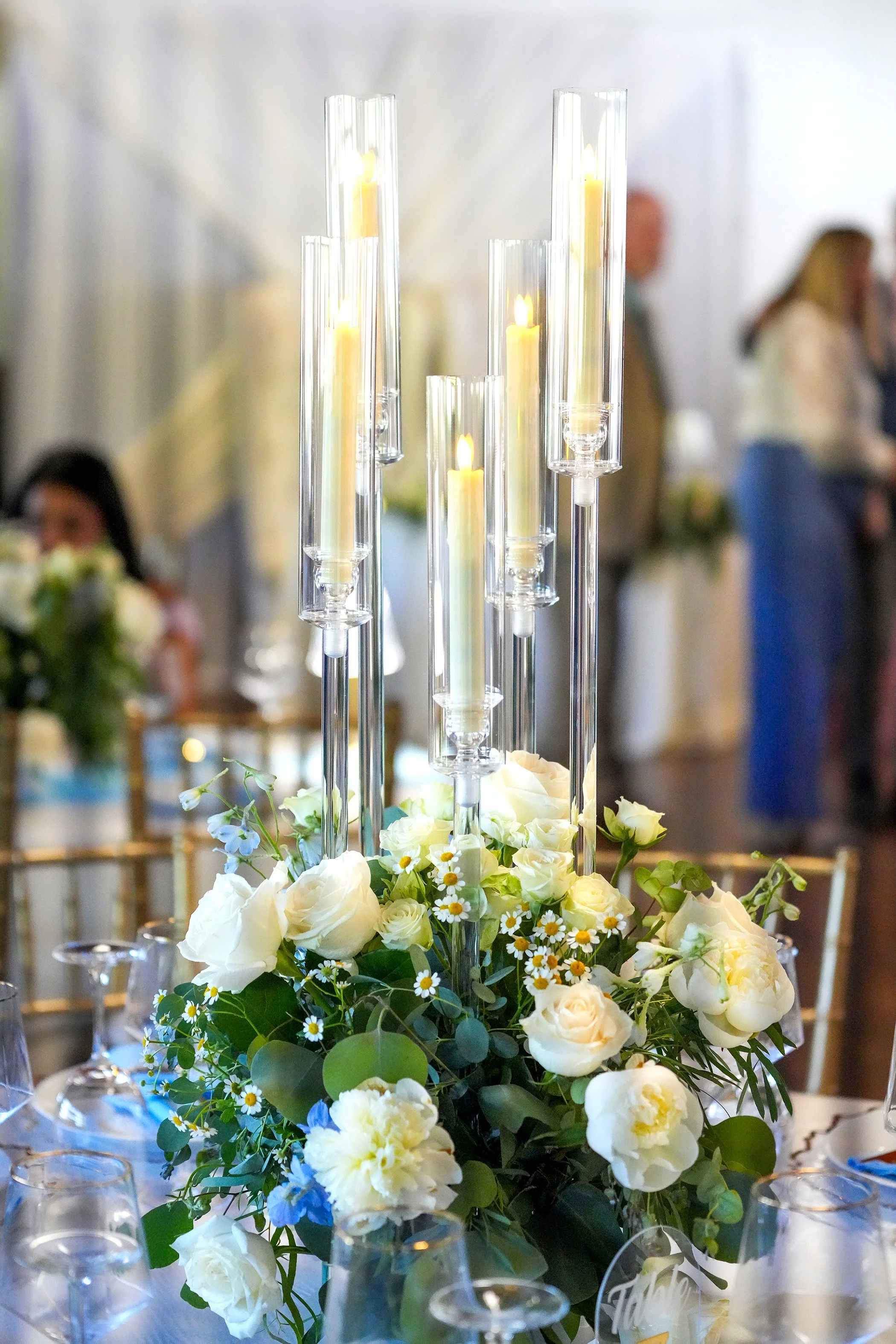Elegant wedding centerpiece featuring white roses, peonies, and tall glass candle holders on a reception table.

