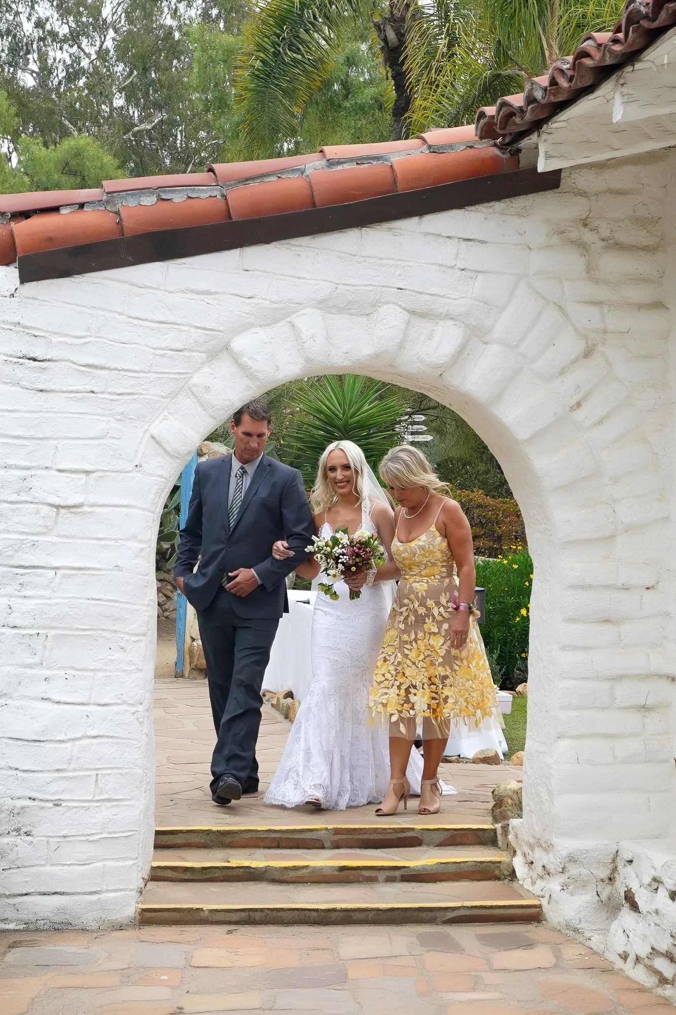 A photo of a bride in a white lace dress and veil walking down an outdoor aisle through a white stucco archway, escorted by a man in a dark suit and a woman in a yellow floral dress in Carlsbad.