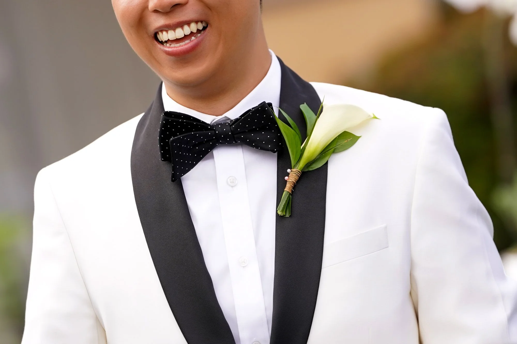 A close-up shot of a groom smiling broadly in a white tuxedo jacket with black lapels, a polka-dot bow tie, and a white calla lily boutonniere.


