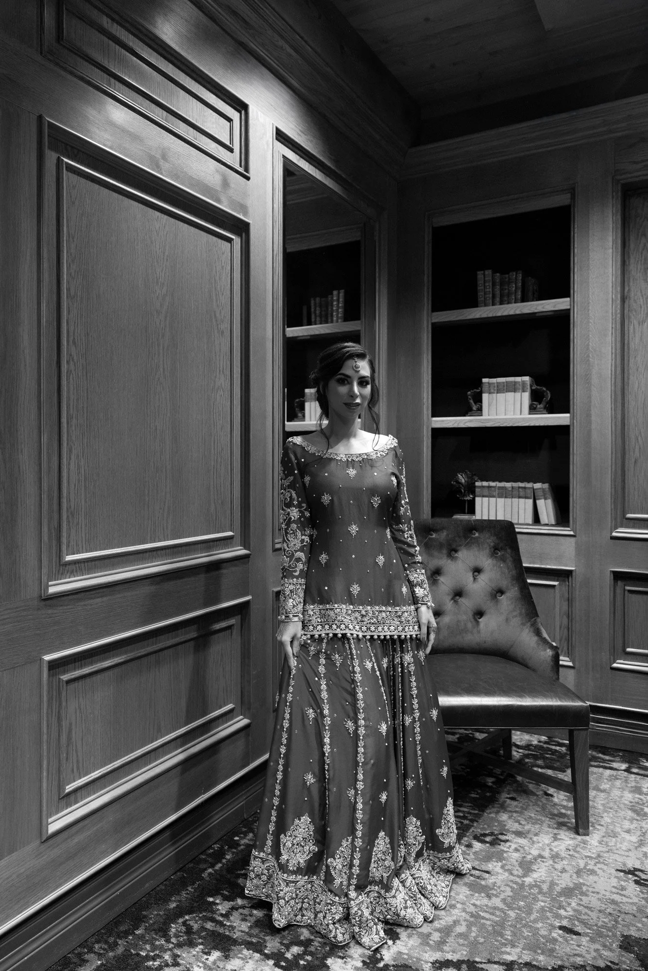 A black and white photograph of a woman in an ornate, traditional Indian or Pakistani formal outfit standing in a room with wood-paneled walls and built-in bookshelves. She is wearing a long-sleeved top (kameez) and a flowing skirt (gharara or leheng