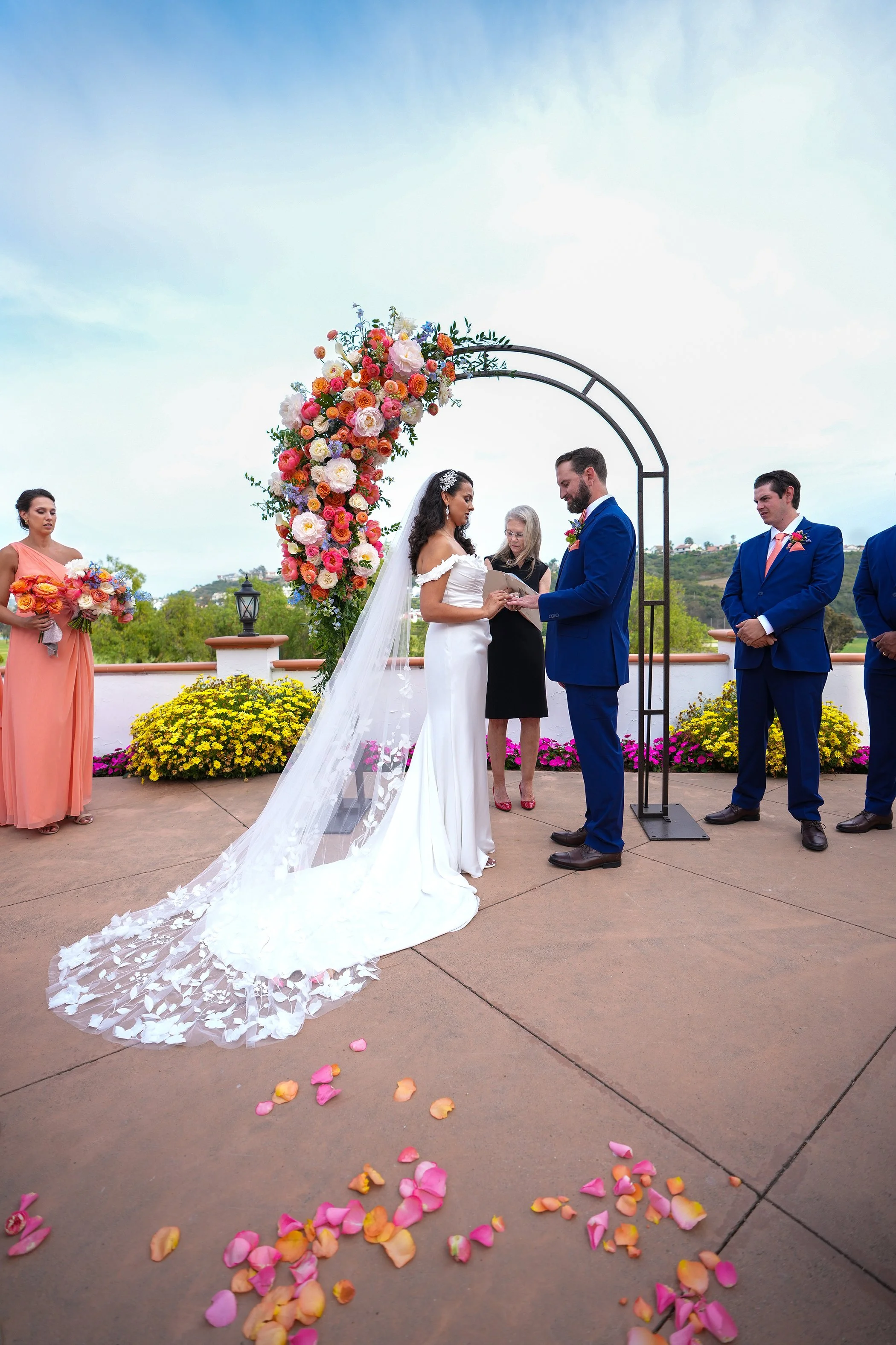 A wedding ceremony photograph by Reimo Photography capturing a bride and groom exchanging rings under a black metal arch adorned with vibrant orange, pink, and white flowers. Pink and orange rose petals are scattered on the patio in the foreground, w