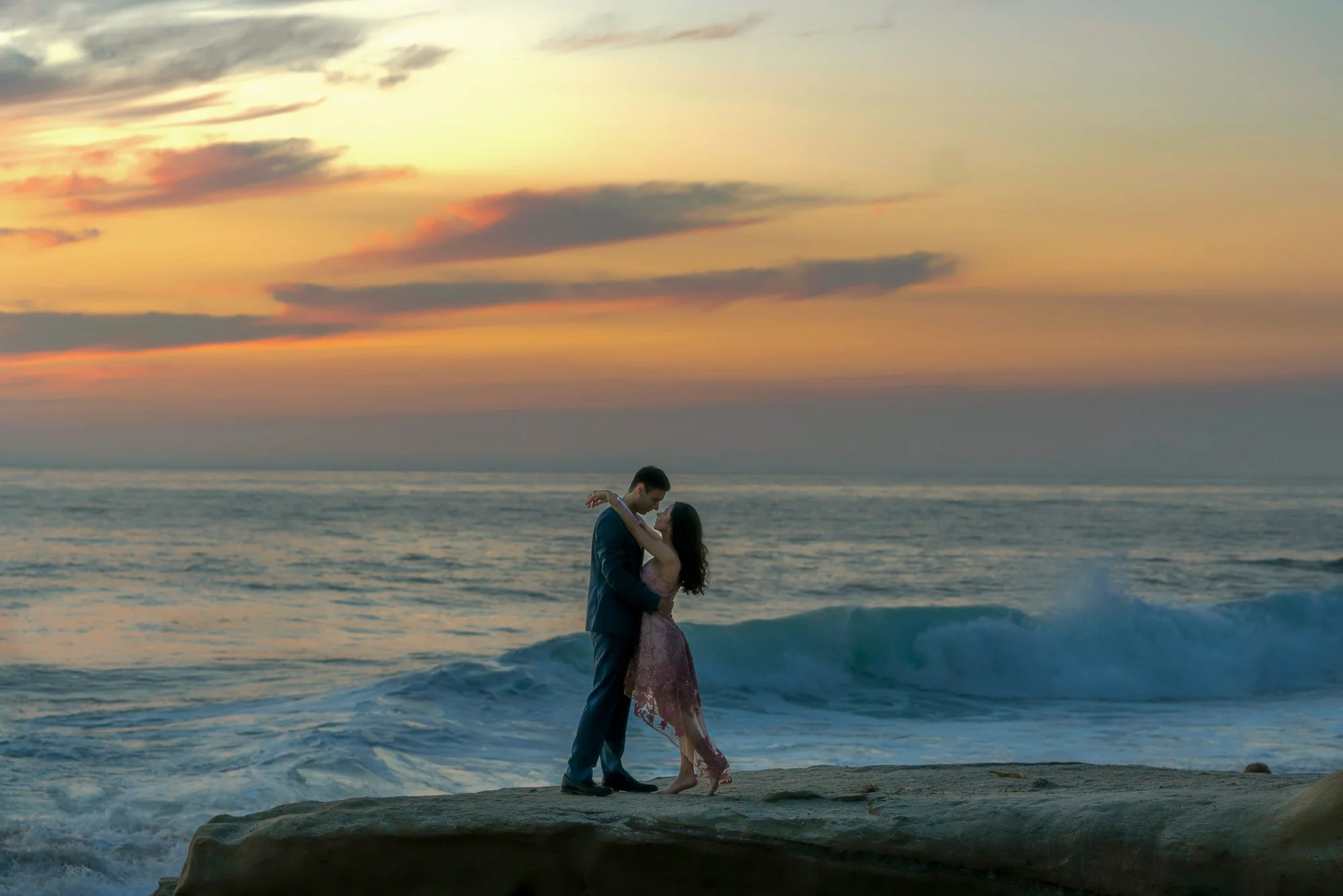 An outdoor photograph of a couple embracing on a large rock formation by the ocean during a beautiful sunset or sunrise. The man is wearing a dark suit, and the woman is in a flowing, patterned dress. The sky is filled with warm orange, pink, and pur